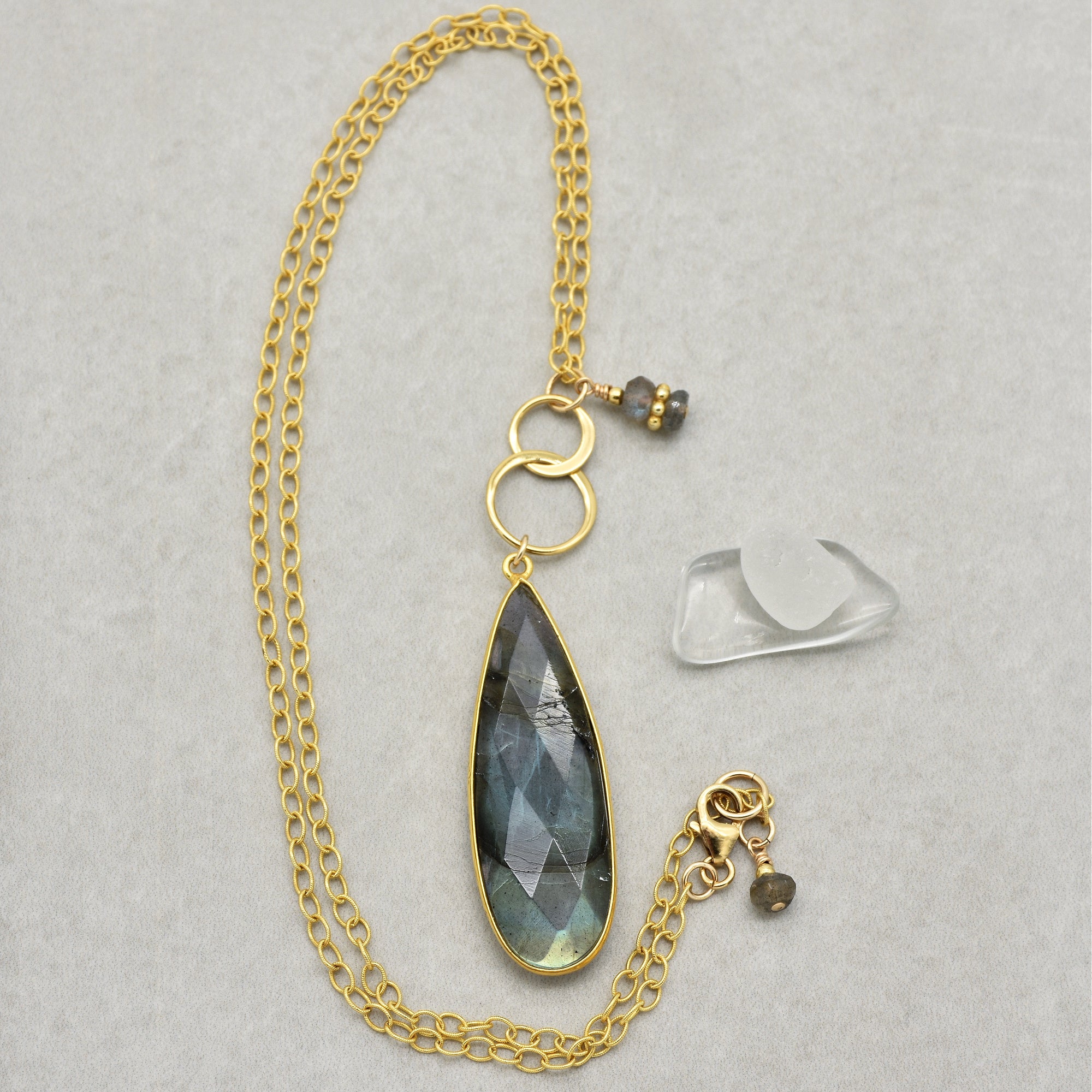This image shows a necklace. It features a gold chain with a large teardrop-shaped pendant that has a faceted gemstone setting. There are also two smaller gold rings and small bead charms attached near the clasp. The overall design and the length of the chain indicate that this piece of jewelry is a necklace.