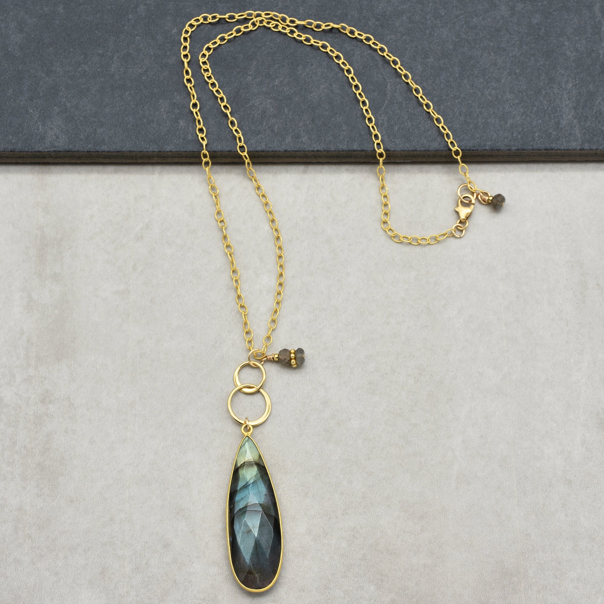 This image shows a necklace. It features a gold chain with a teardrop-shaped pendant that has a faceted dark stone, likely labradorite, hanging from it. There are also two gold rings above the pendant and a small decorative bead near the clasp.