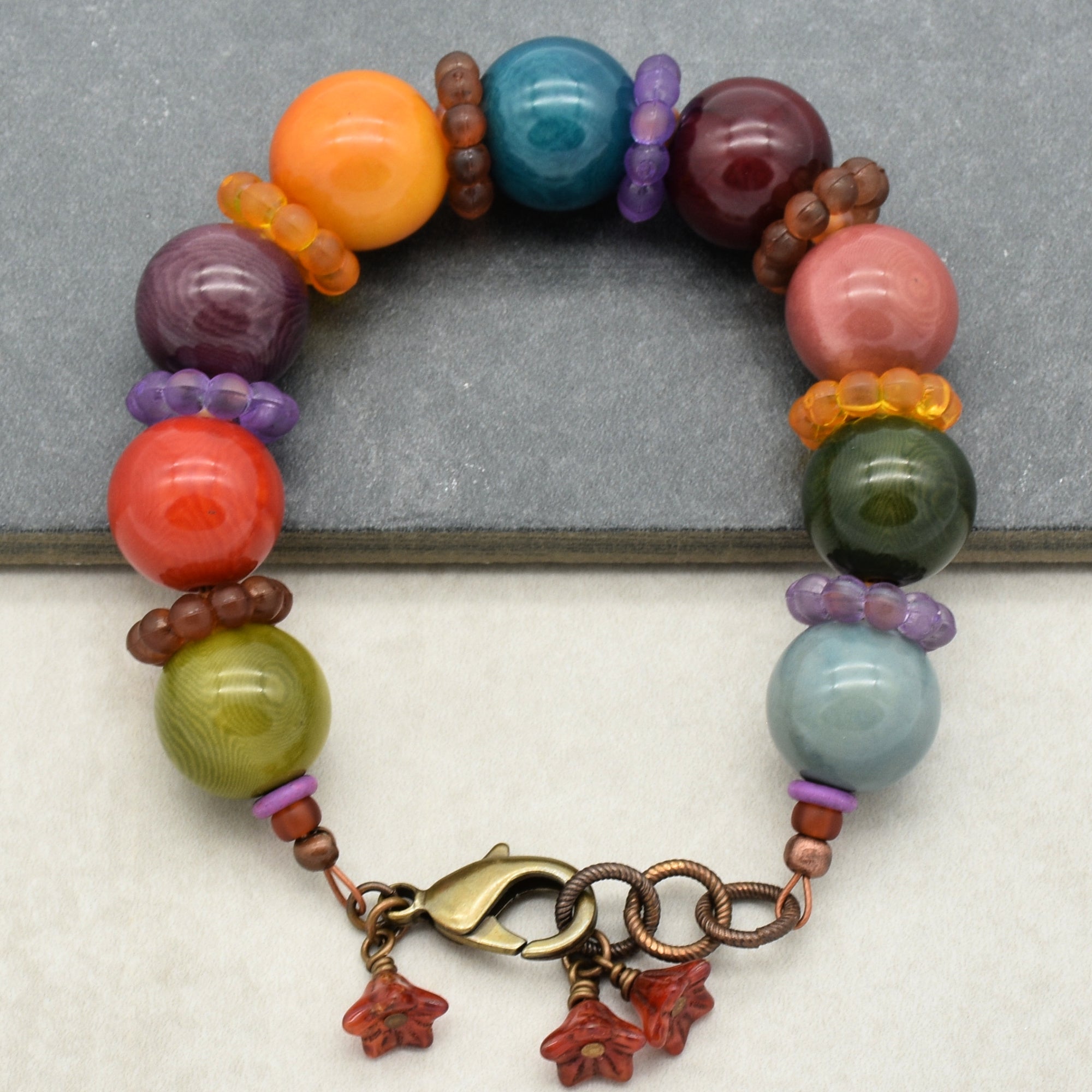 This image shows a bracelet. It is composed of large, colorful beads in various shades, separated by smaller ring-like beads. It has a metal clasp with a few small dangling charms attached. The overall design and size indicate that it is a bracelet.