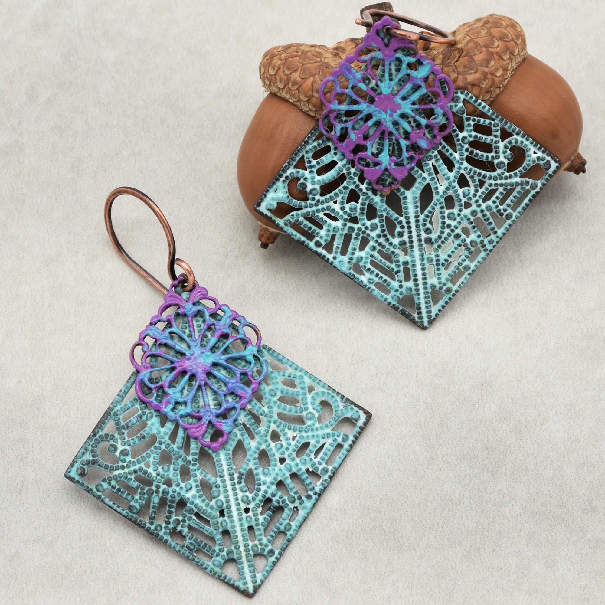 This image shows a pair of earrings. The jewelry has intricate, lacy metal designs with a combination of turquoise and purple colors, and they have hooks for wearing on ears.