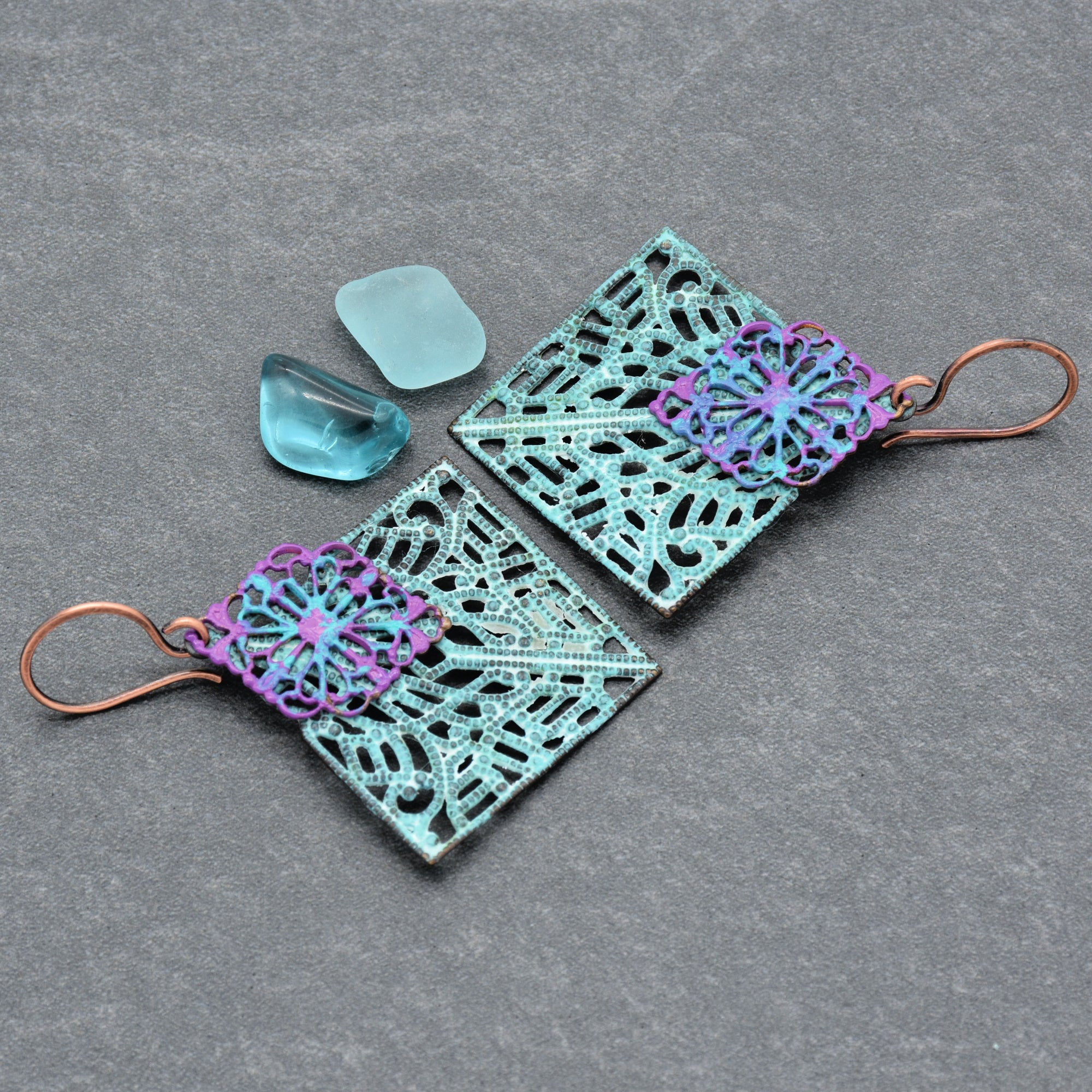 The image shows a pair of earrings. They have a decorative, intricate design with blue and purple elements and copper-colored hooks for wearing on the ears.
