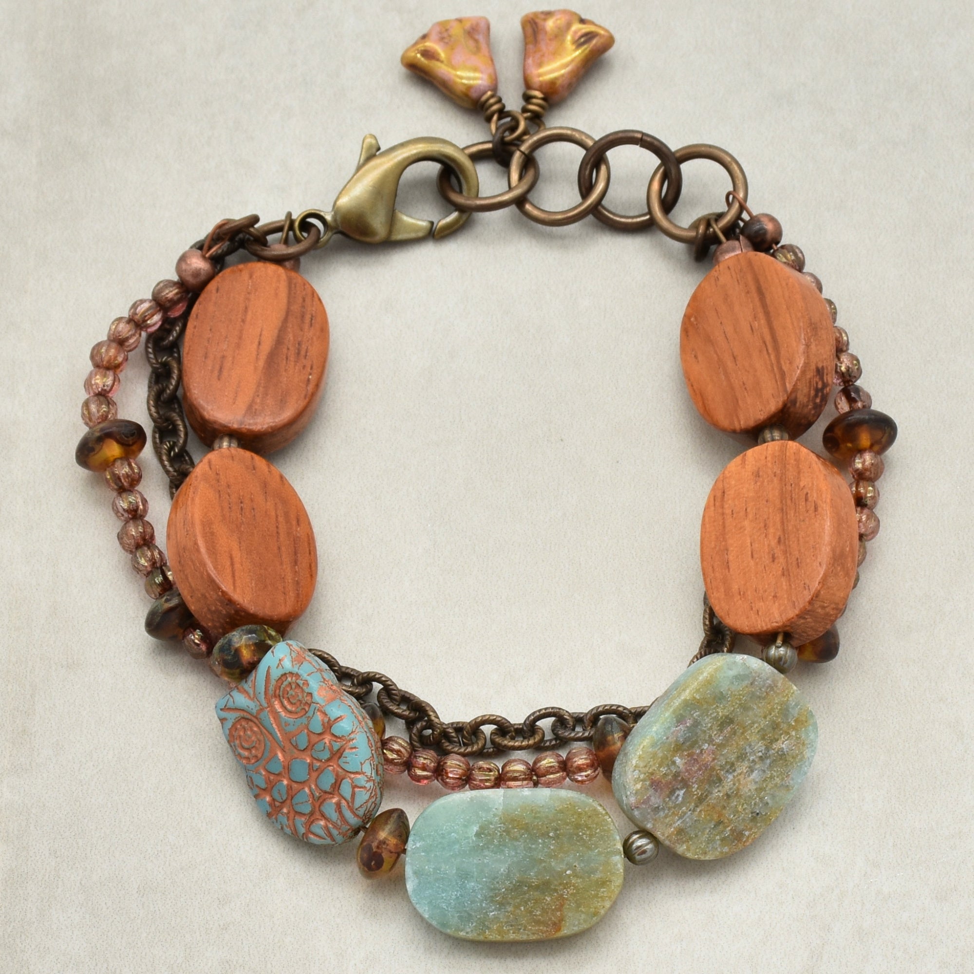 This image shows a bracelet. It features large wooden oval beads, smaller round beads, and a few stones, including one with a turquoise color and decorative patterns. The bracelet has a metal chain and clasp for fastening.