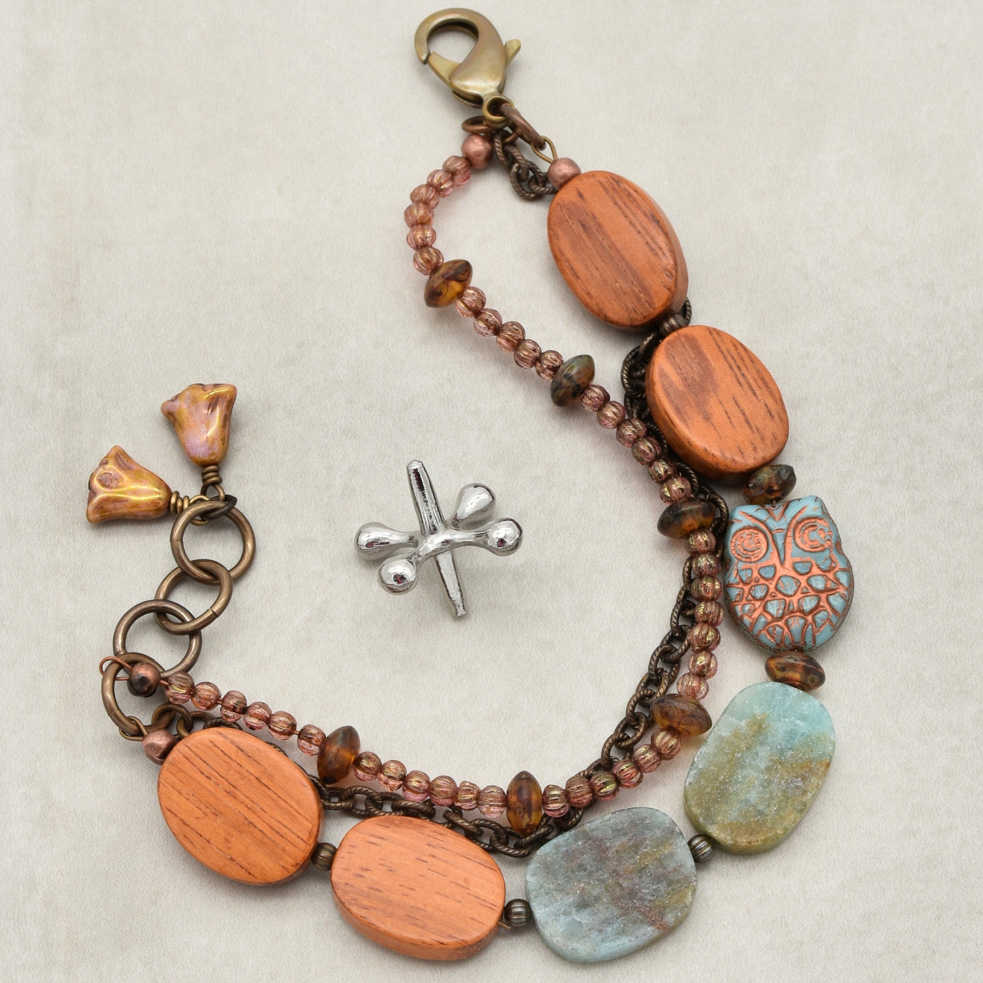 This image shows a bracelet. It has a lobster clasp, various beads including wooden ones, some blue-green stones, and a decorative bead shaped like an owl. The size and shape of the piece indicate that it is meant to be worn around the wrist.