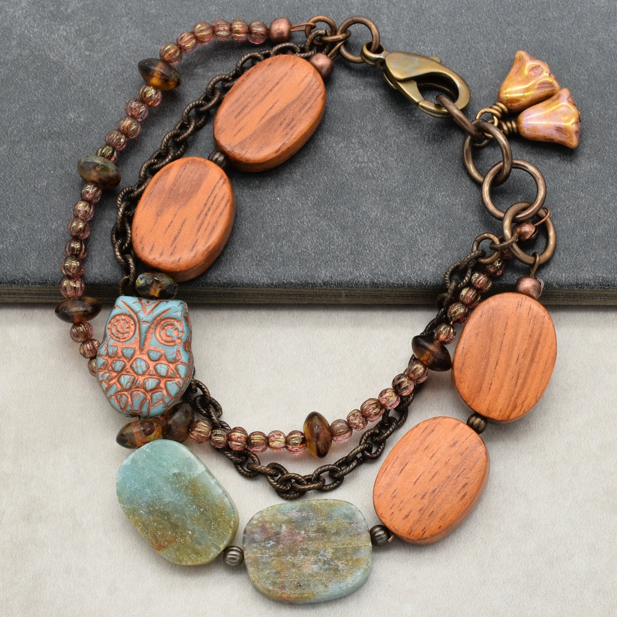 This image shows a bracelet. It has a clasp for fastening and features various beads, including wooden oval beads, a blue owl-shaped bead, and some greenish stone-like beads, along with chains and smaller accent beads.