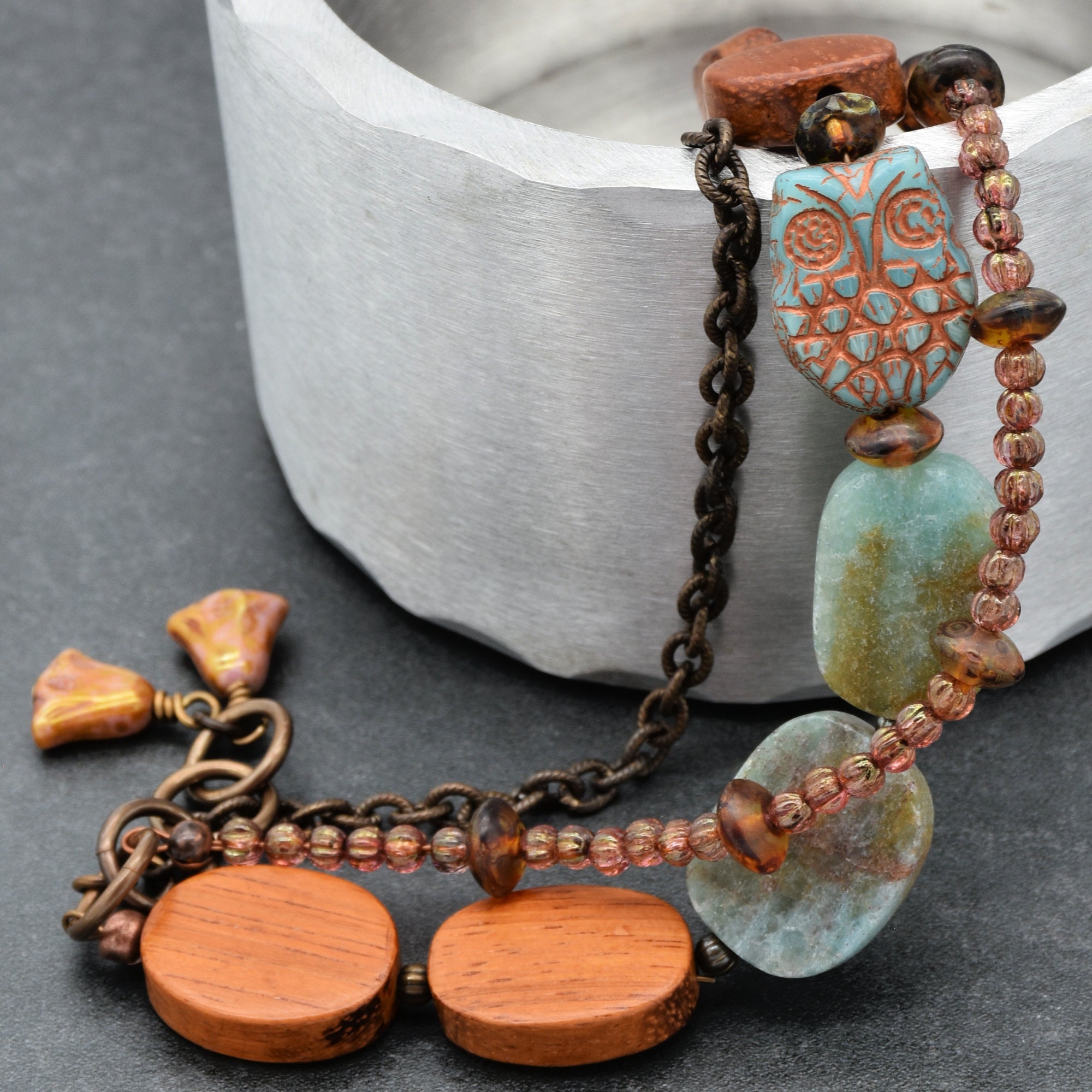 The image shows a bracelet. It features a combination of materials including a chain, wooden beads, glass beads, and a carved turquoise owl-shaped bead. The size and design indicate it is meant to be worn around the wrist.
