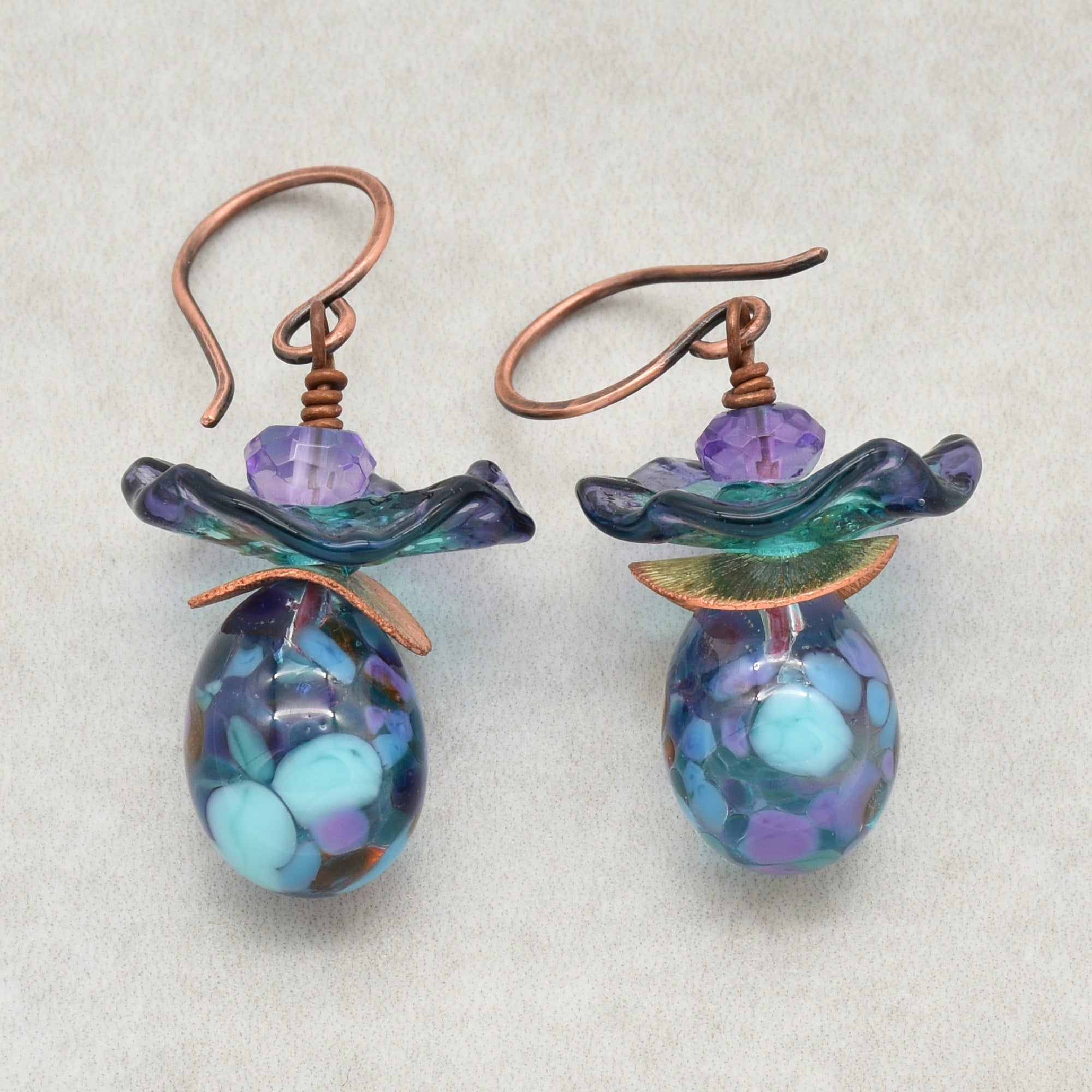 The image shows a pair of earrings. They have hooks for ear piercing and decorative elements featuring purple and blue beads with metallic accents.