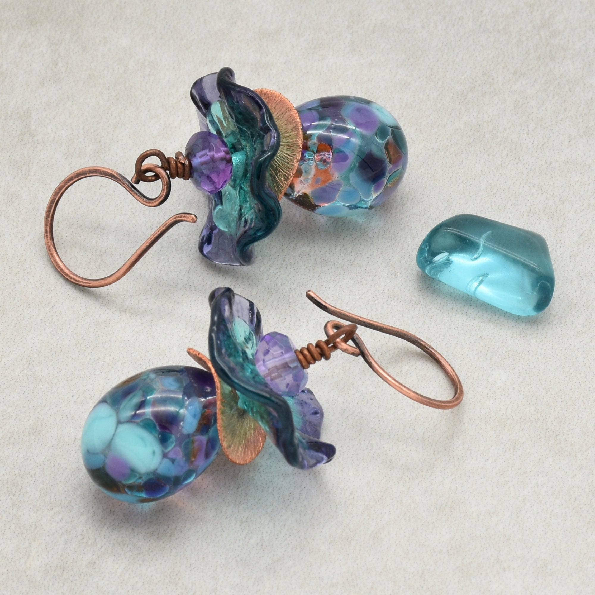 The image shows a pair of earrings. The jewelry pieces have hooks that are characteristic of earrings, and the decorative parts include colorful, intricate glass beads with copper wire wraps.