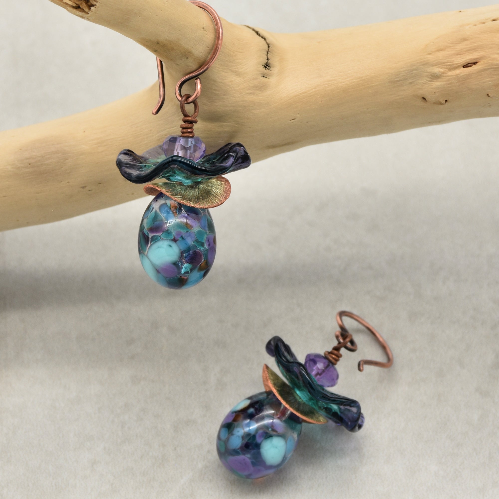 The image shows a pair of earrings. They have a hook-style attachment and decorative elements including a purple and blue teardrop-shaped bead, a wavy glass piece, and a small copper accent.