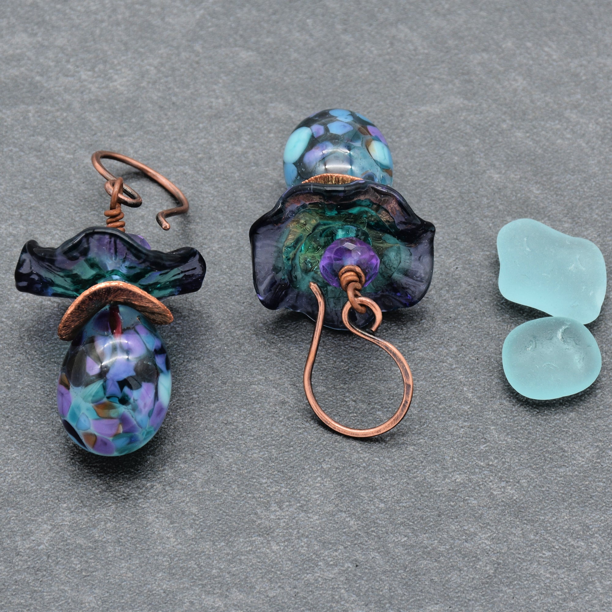 The image shows a pair of earrings. They have a decorative design with beads in blue, purple, and teal colors, and copper wire hooks for wearing on the ears.