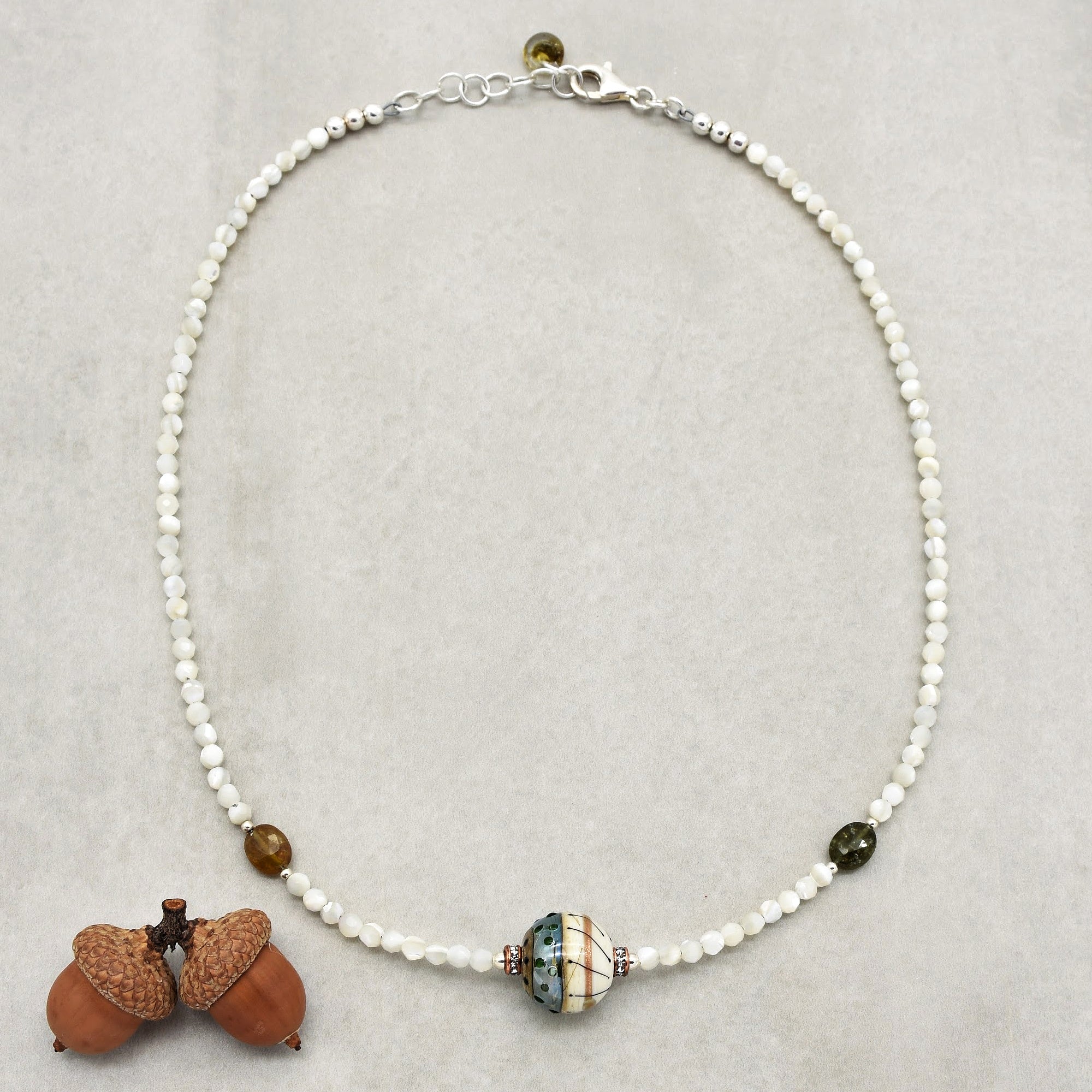 This image shows a necklace. It is composed of small white beads with a larger decorative bead in the center and two smaller colored beads on either side. The clasp and chain extender at the top confirm that it is a necklace.