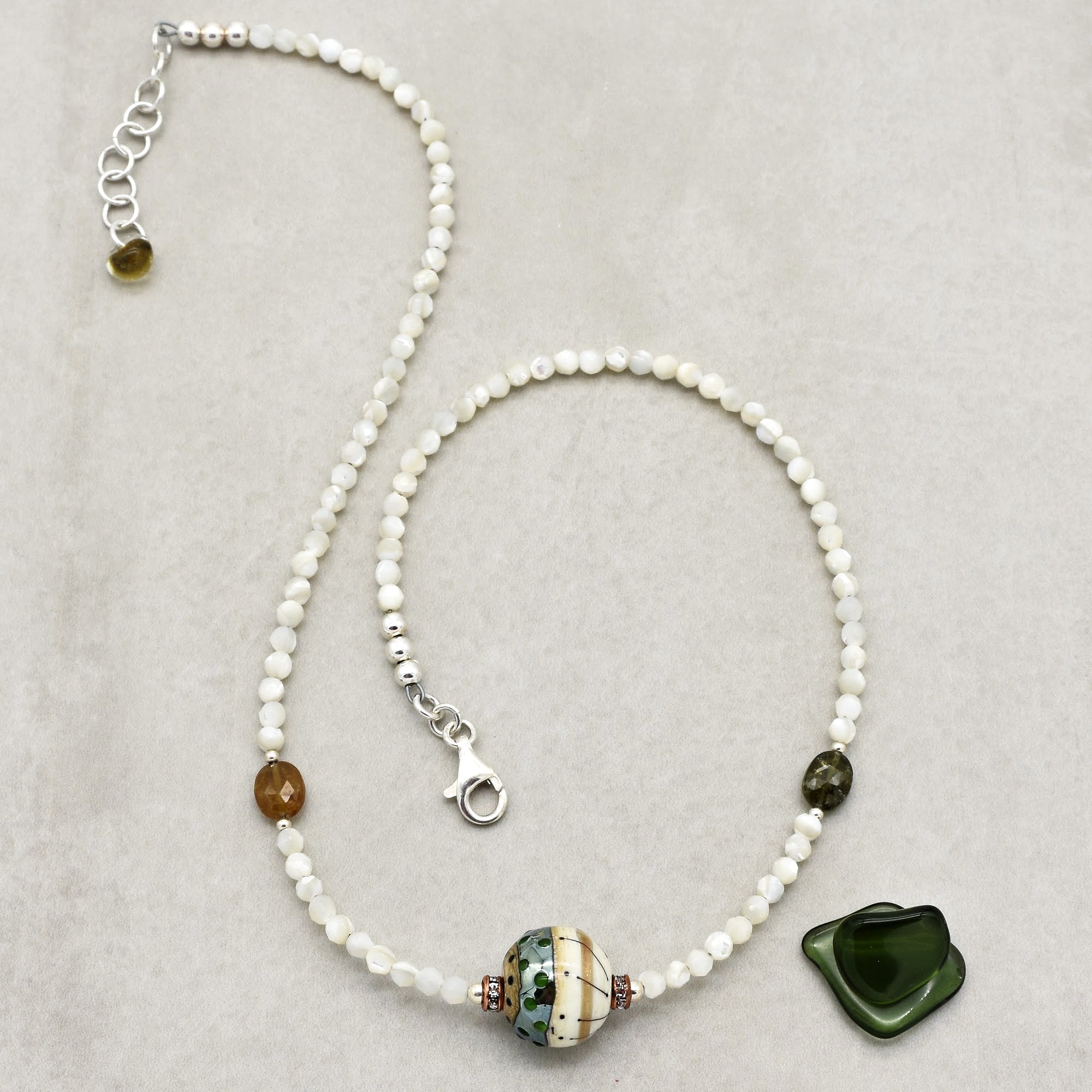 The image shows a necklace. It consists of small white beads strung together, with a larger decorative bead and a green pendant as focal points. The necklace has a clasp for fastening.