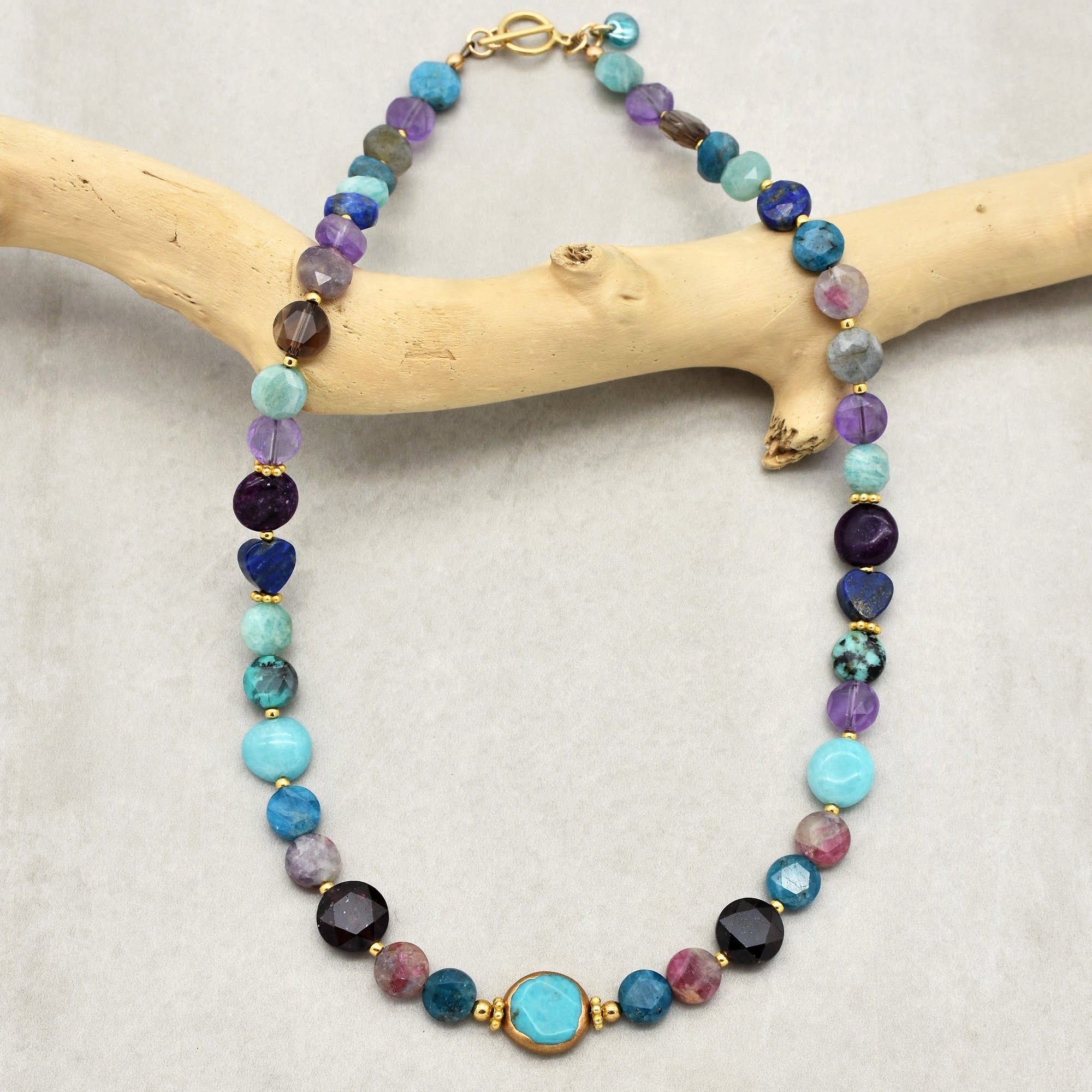 This image shows a necklace made of various round and faceted beads in different colors, including shades of blue, purple, and pink, with some gold accents. It has a gold clasp at the top.