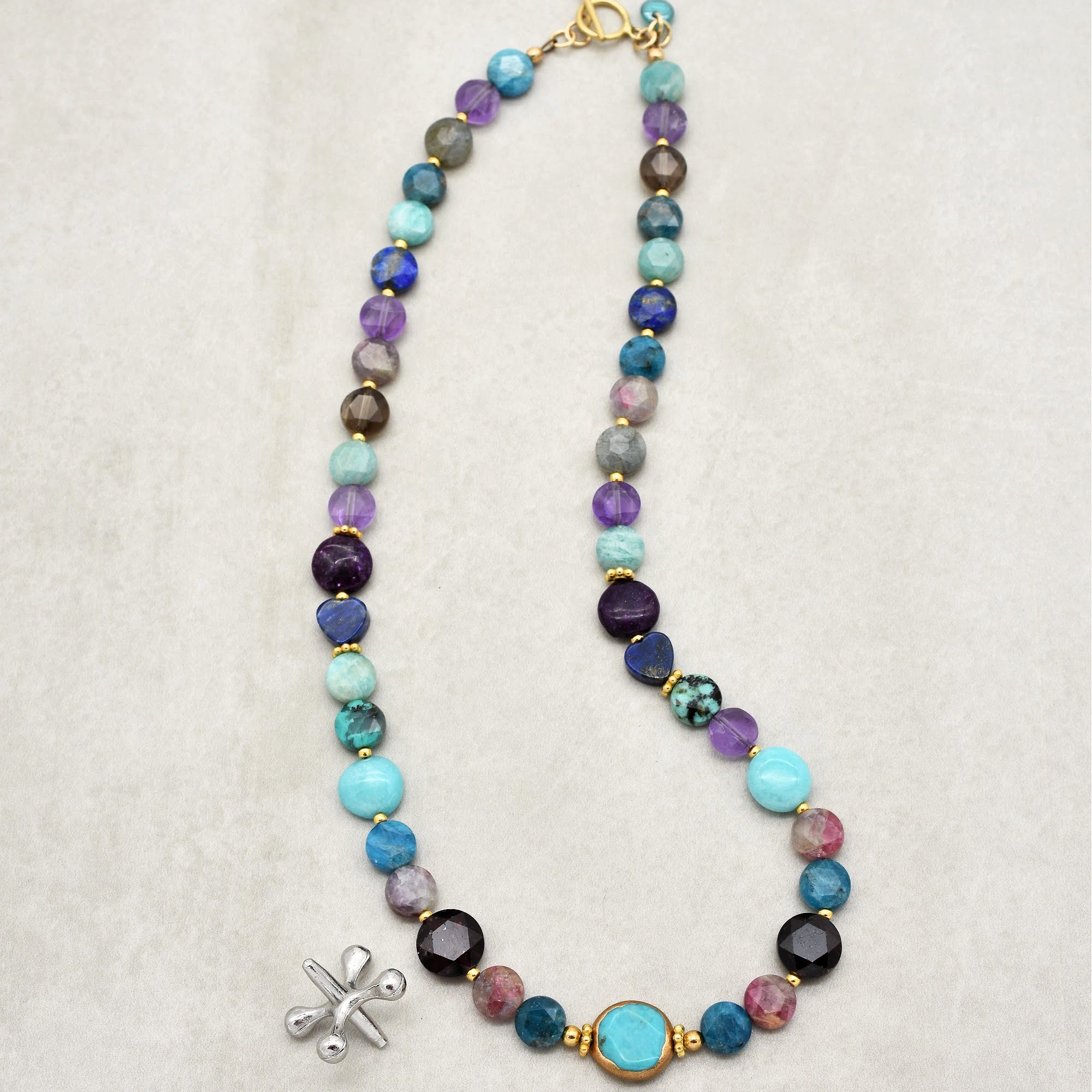 This image shows a necklace. The jewelry piece has a chain of variously colored and shaped beads, including turquoise, purple, blues, and gold spacers, with a clasp at the top for fastening around the neck. It is too long and designed to be worn around the neck, so it is not an earring or bracelet.