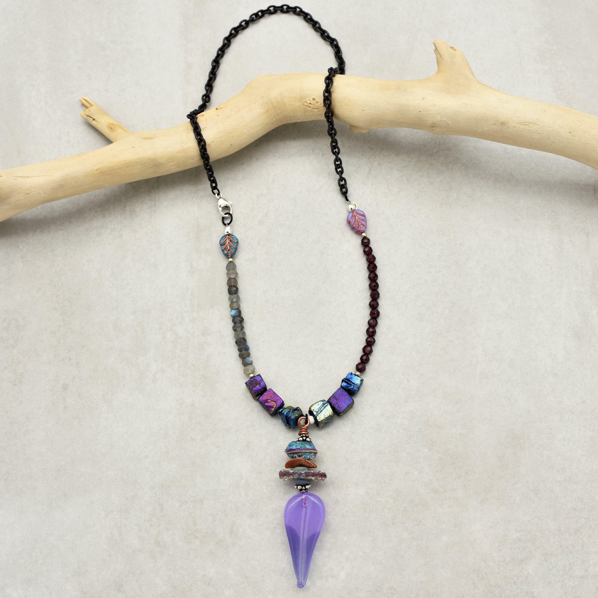 This image shows a necklace. It features a combination of a black chain and various beads, with a purple teardrop-shaped pendant hanging at the center.