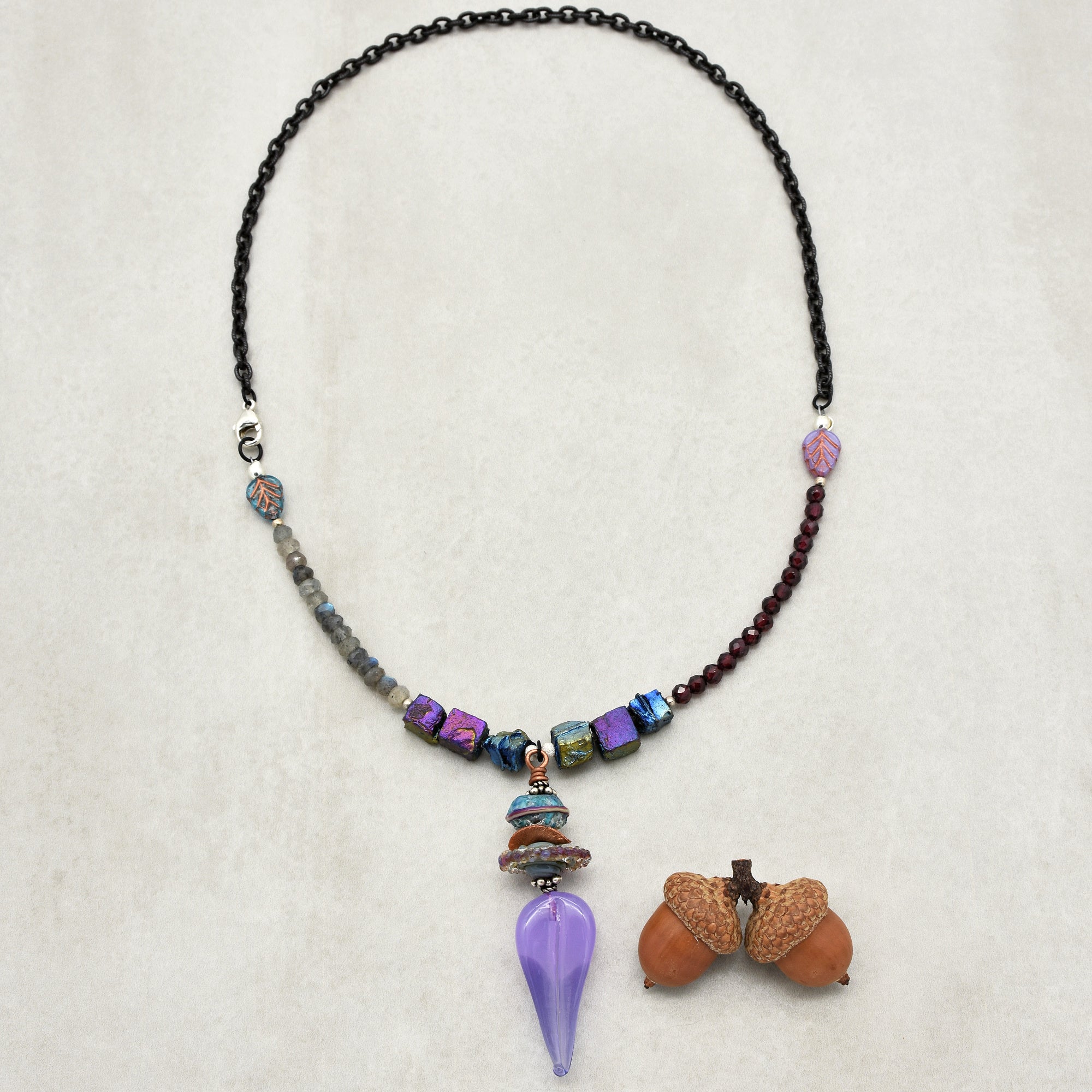 This image shows a necklace. It has a black chain with a section of different beads, including purple, blue, and gray beads, and a distinctive purple pendant hanging in the center. There are also two acorns placed beside the necklace.