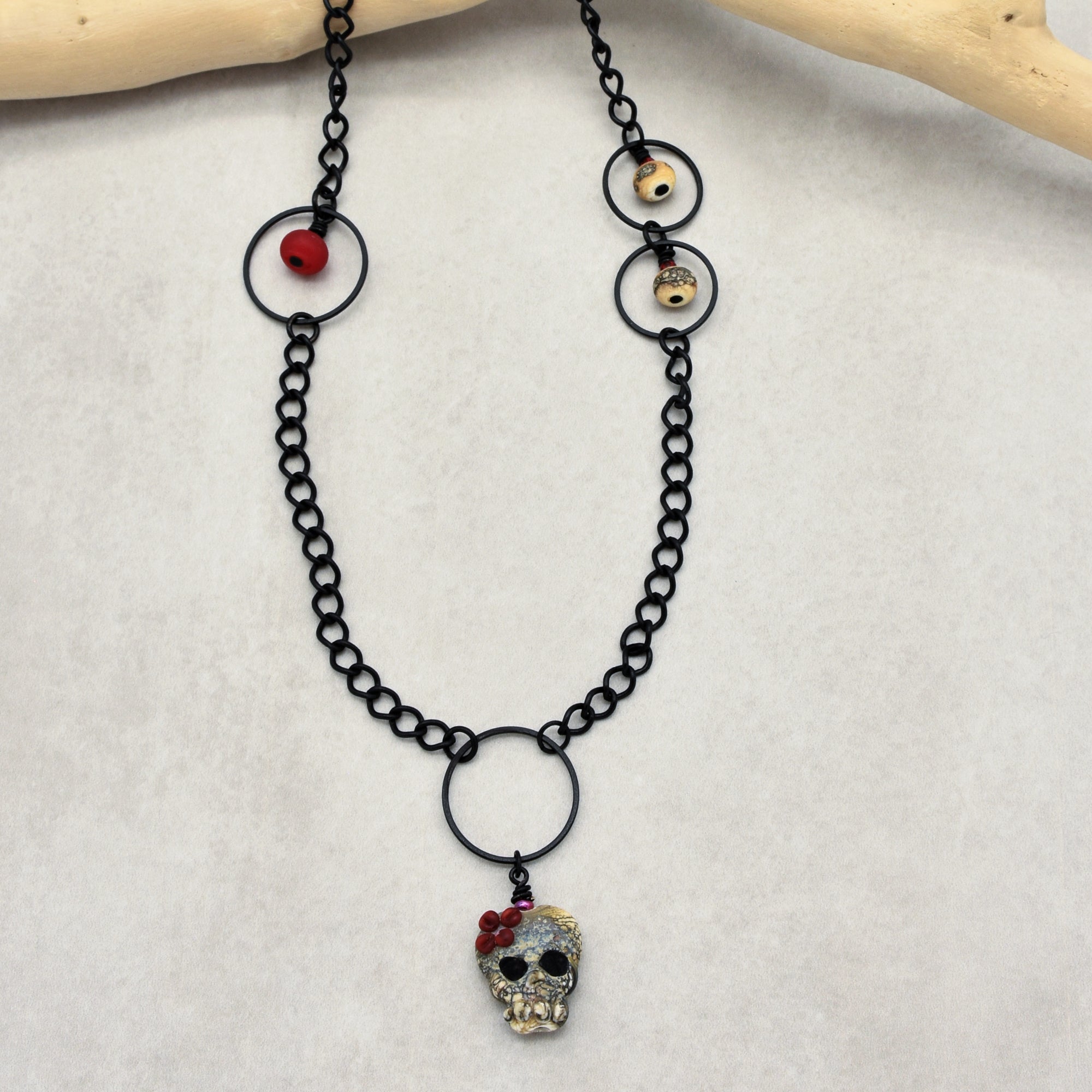 This image shows a necklace. The black chain features a skull pendant adorned with red beads, and there are additional decorative beads attached to the chain.