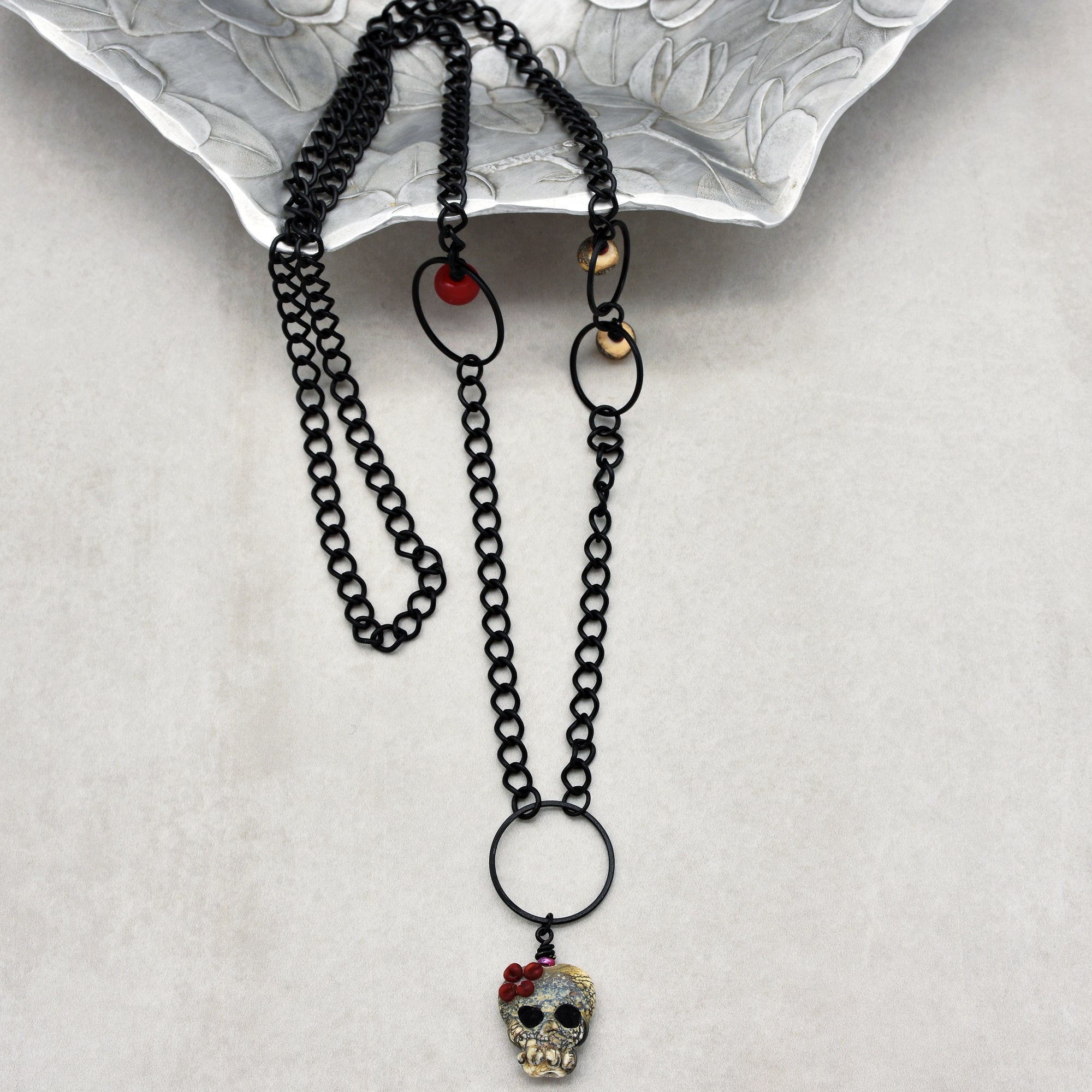 This image shows a necklace. The jewelry piece has a black chain with a skull pendant attached, decorated with small red beads. The length and design suggest it is meant to be worn around the neck.