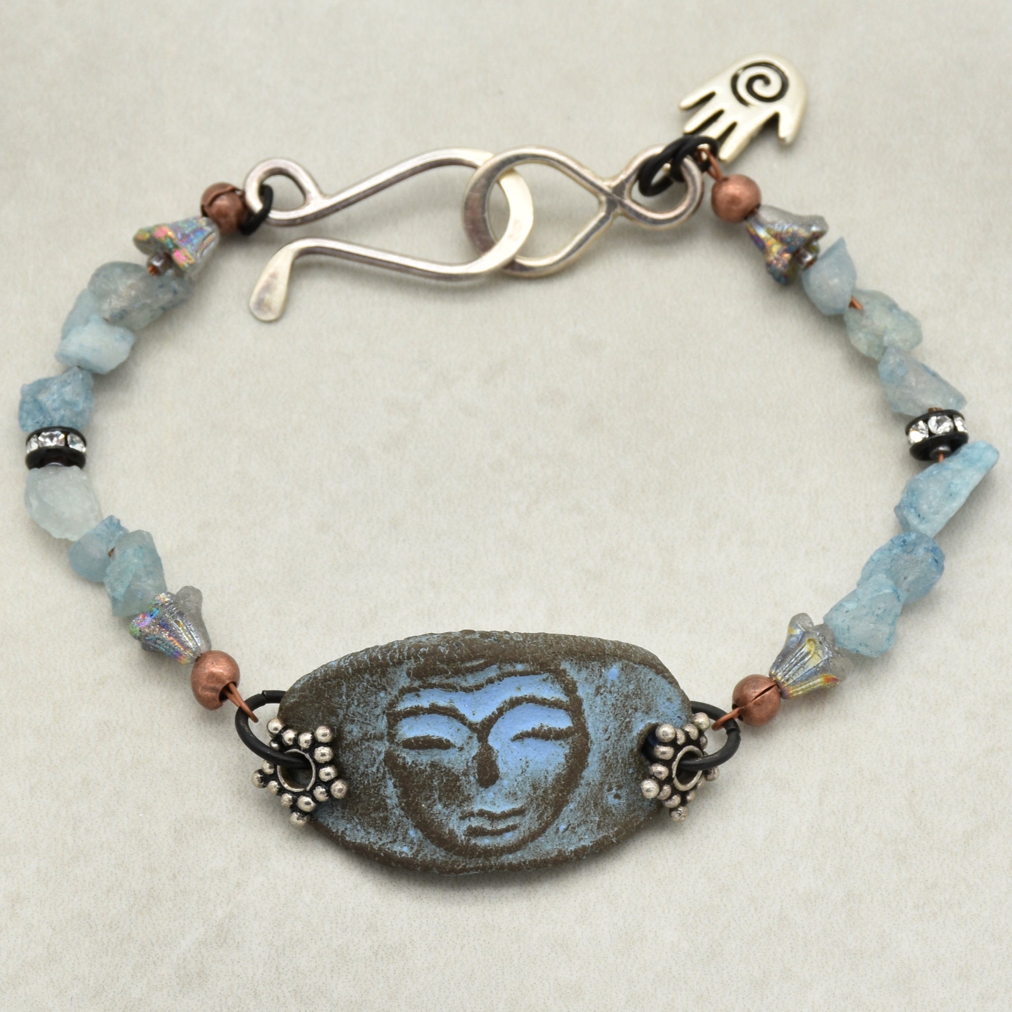 This image shows a bracelet. It features a central rectangular piece with a face design, flanked by small decorative beads and blue stone-like beads. The bracelet has a unique hook-and-eye clasp at the top.