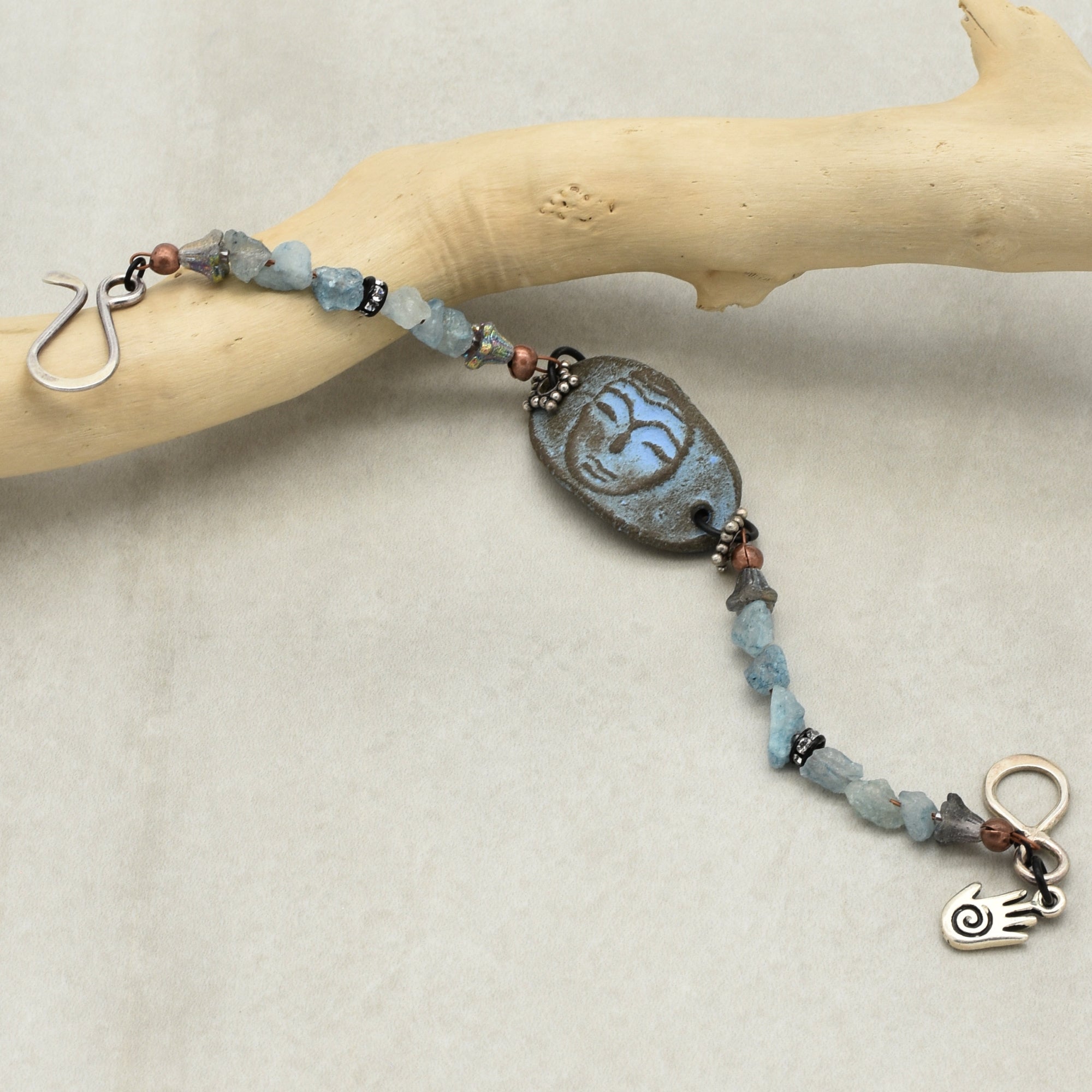 This image shows a bracelet. It features an artistic central bead with a face design, flanked by blue stone beads and other decorative elements. The clasp and size also indicate that it is meant to be worn around the wrist.