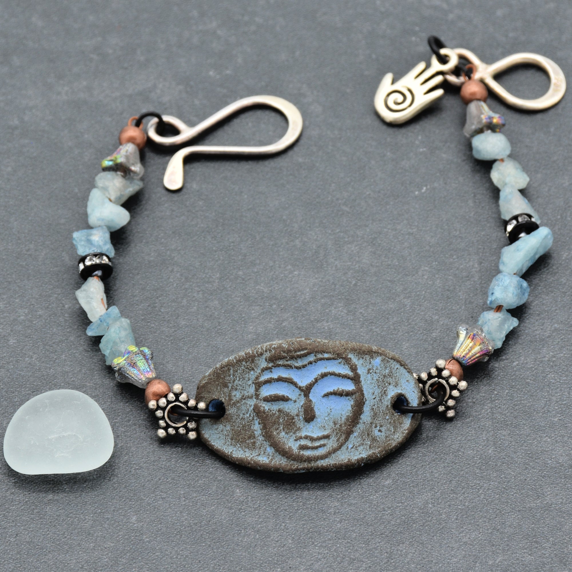 The image shows a bracelet. It features a central oval piece with a rustic finish, surrounded by small decorative beads and blue stone chunks on either side. The bracelet has a unique hook clasp and a small charm resembling a hand near the clasp.