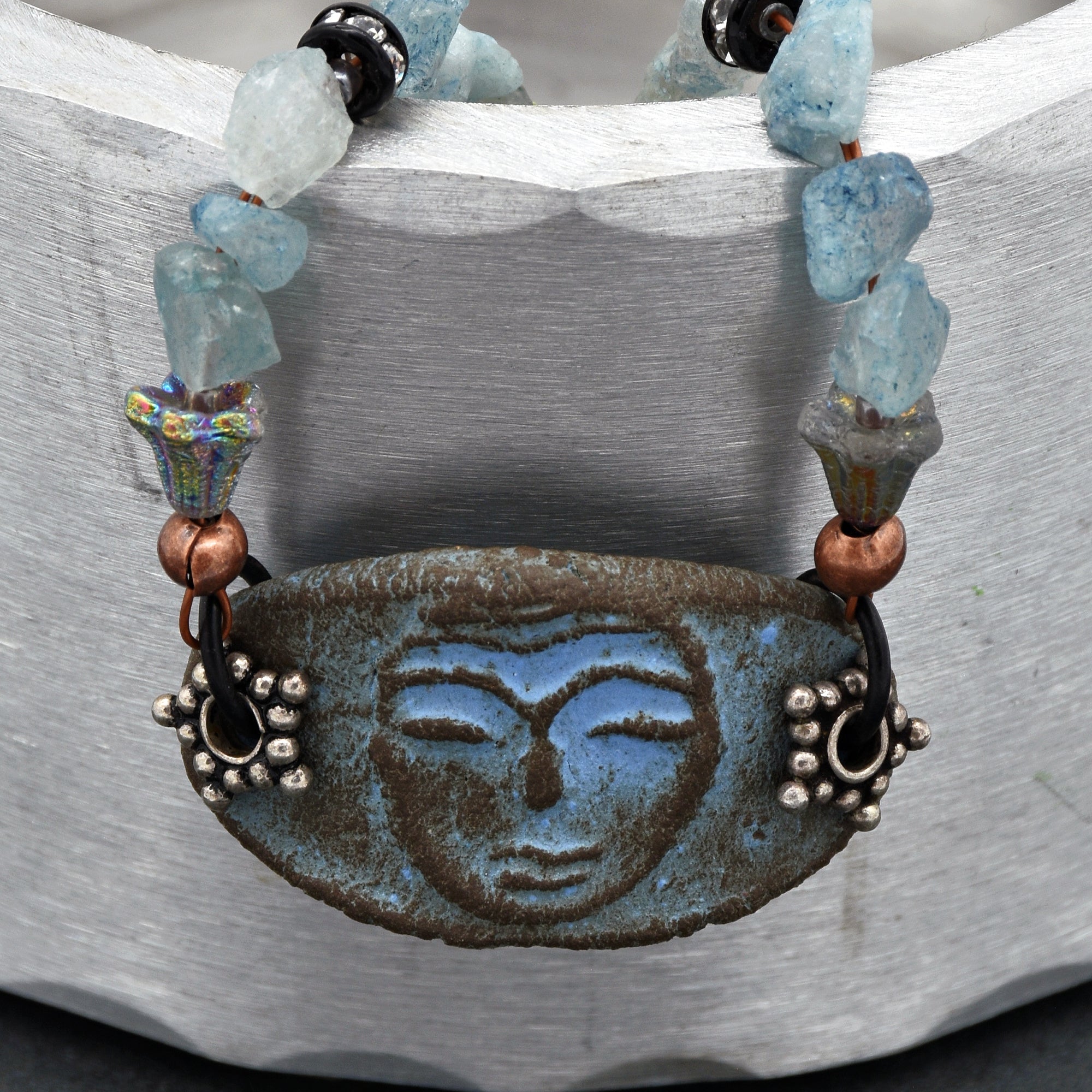 This image shows a piece of jewelry that appears to be a necklace. The focal point is a ceramic or stone pendant with a stylized face design, attached to a string or wire with various beads, including some light blue and iridescent ones. The size and style of the pendant, along with the way it is strung, suggest it is meant to be worn around the neck.