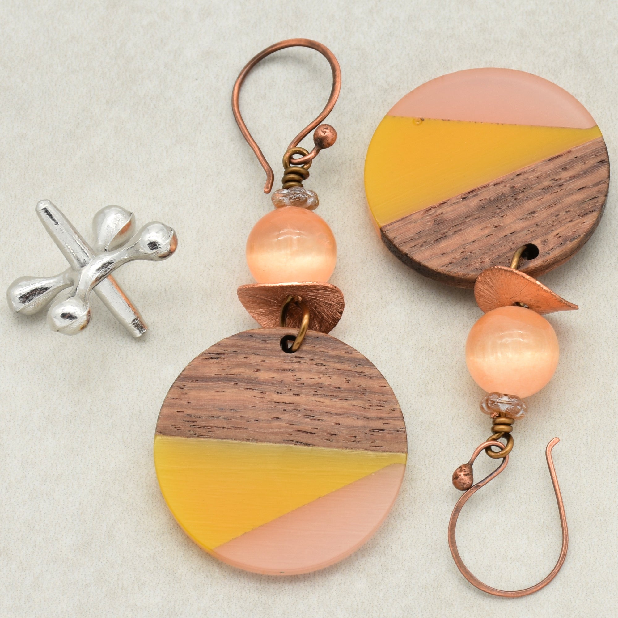 The image shows a pair of earrings. They have a circular wooden component with yellow and pink accents, as well as a round peach-colored bead and a copper or bronze-colored hook for wearing on the ear. There is also a separate silver pin-like piece which is not a part of the earrings.