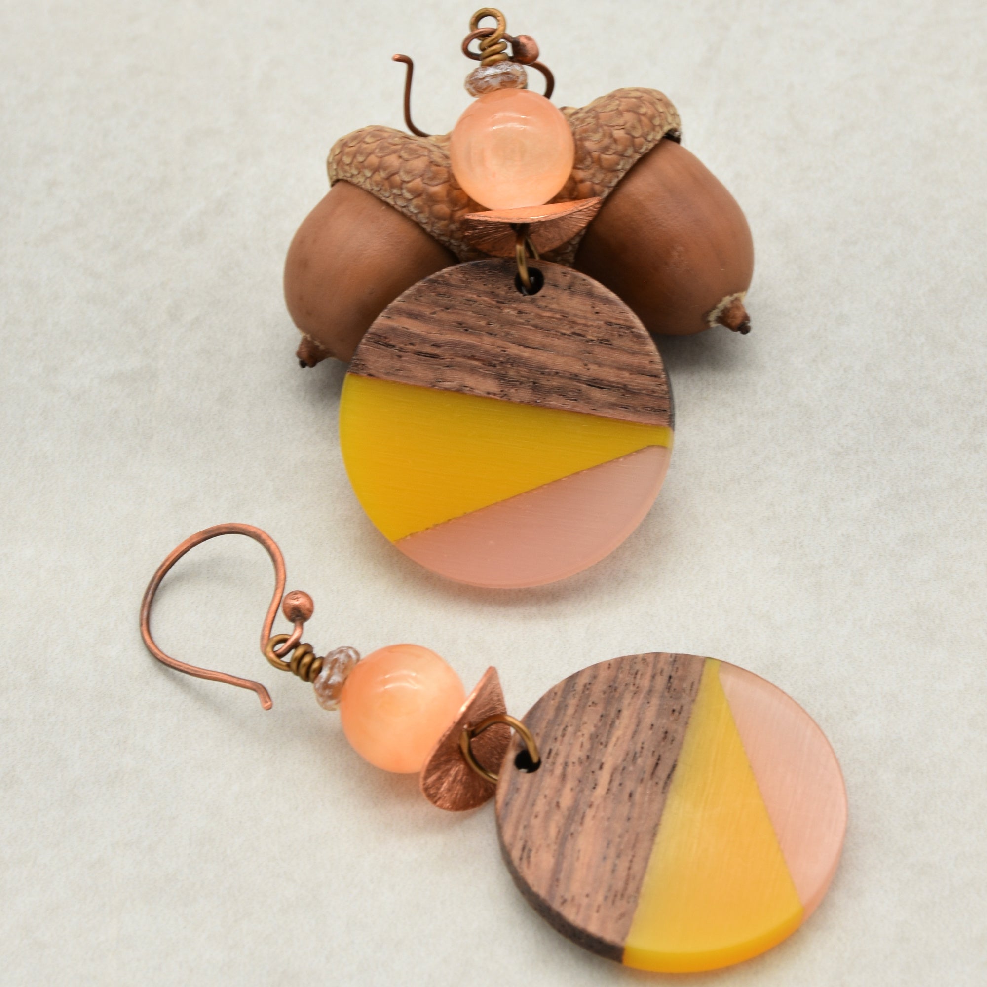 The image shows a pair of earrings. They have a round, multicolored pendant featuring wood, yellow, and pink sections, with a round peach-colored bead above the pendant and a copper-colored hook for wearing.