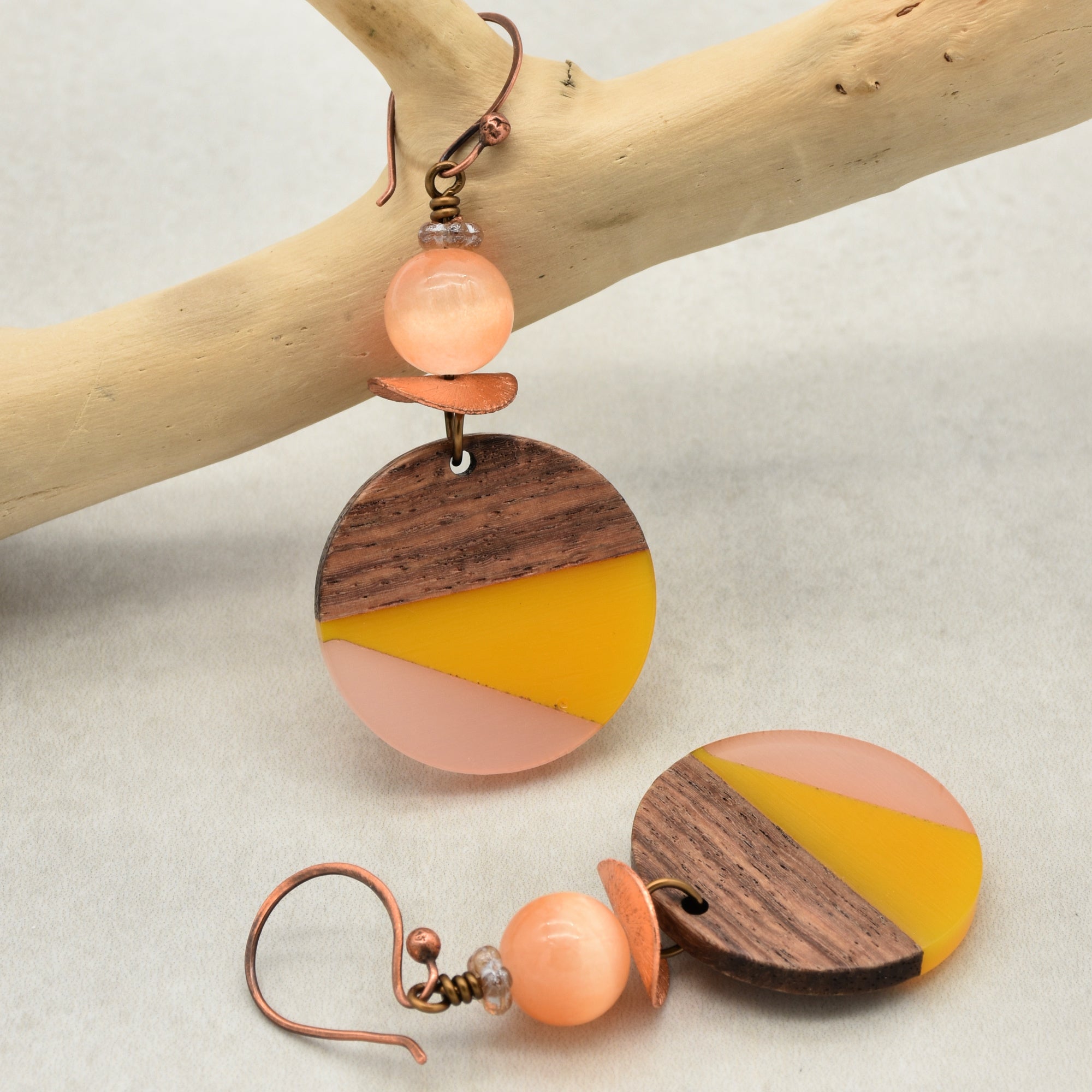 The image shows a pair of earrings. They have a round, color-block design made of wood and resin in brown, yellow, and peach colors, with a peach-colored bead and copper hooks for wearing on the ears.