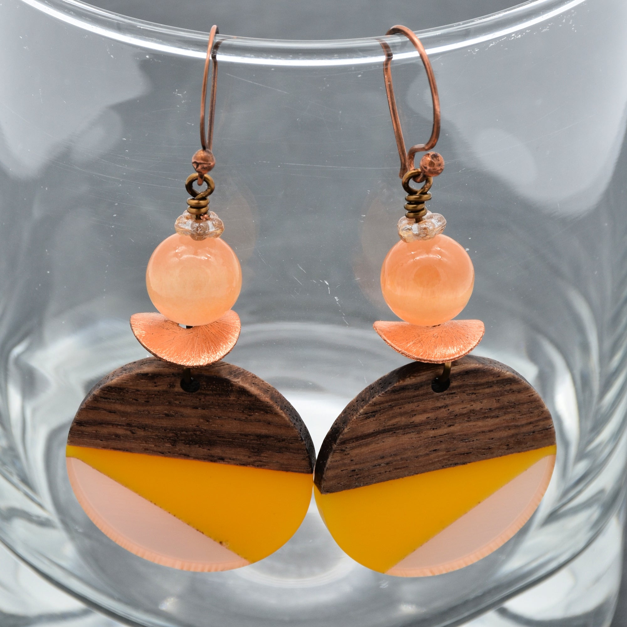The image shows a pair of earrings. They have a hook for the ear, a round peach-colored bead, a small copper detail, and a larger circular piece that is half wood and half a yellow and light pink geometric pattern.