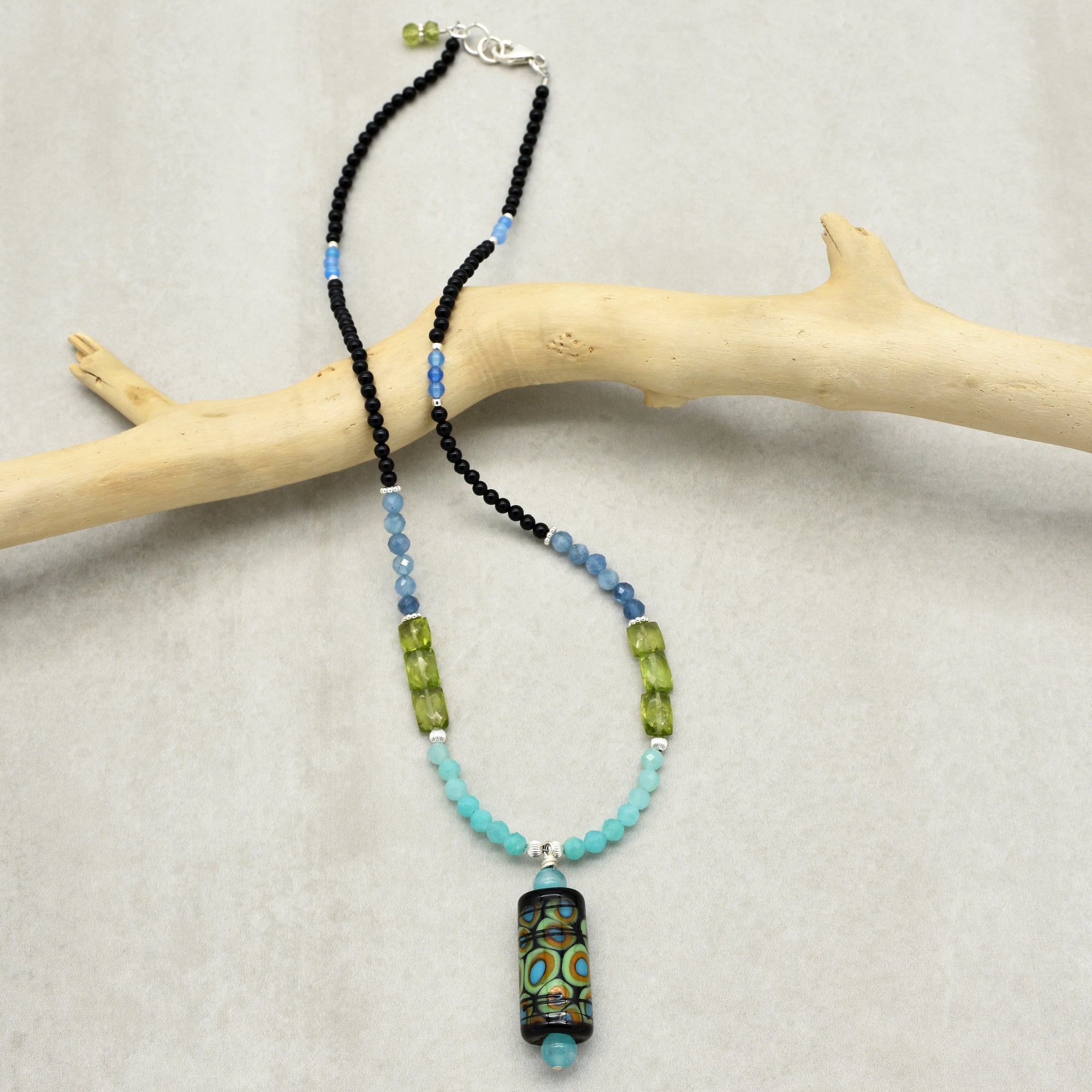 The image shows a necklace. It features a variety of beads in black, blue, green, and turquoise shades, with a decorative cylindrical pendant that has a colorful pattern. The necklace is displayed draped over a light-colored piece of wood.