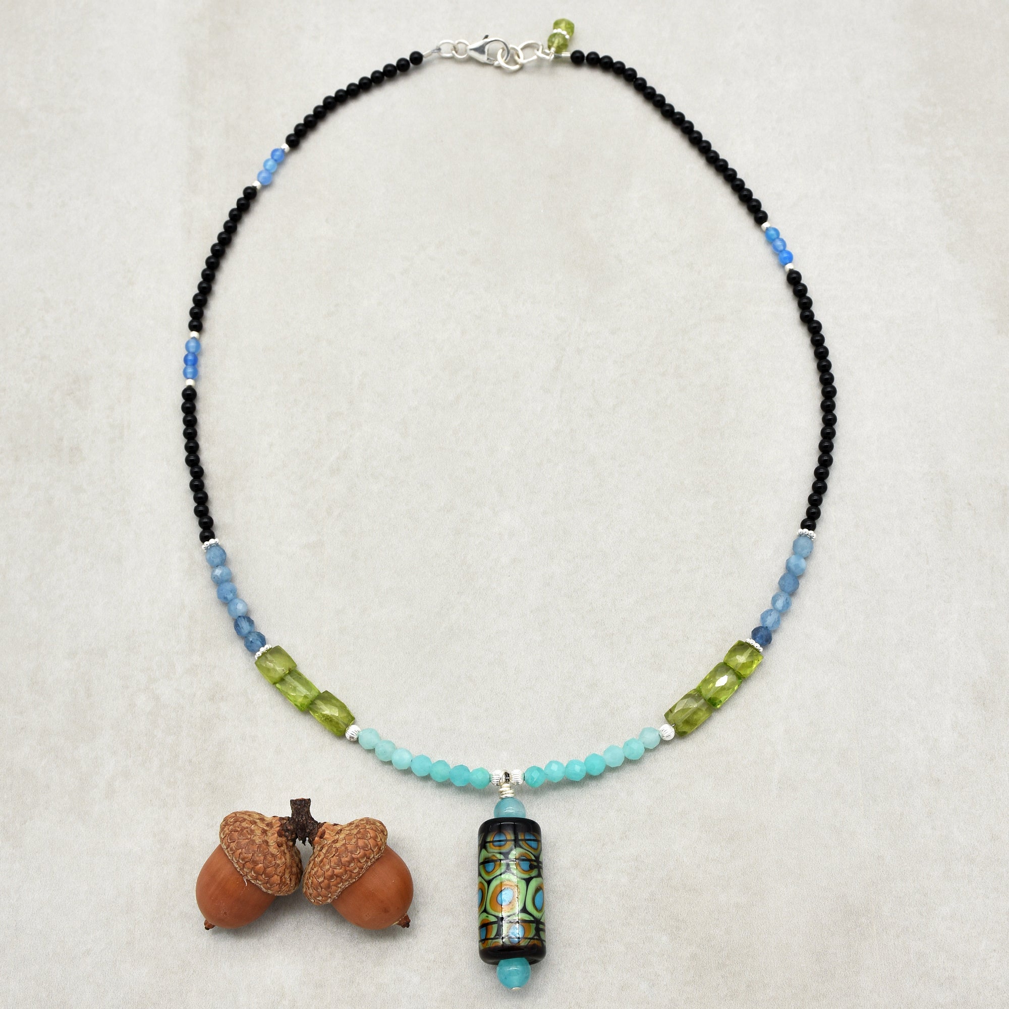 This image shows a necklace. It features a variety of beads in different colors (black, blue, aqua, and green) and a central pendant with a multicolored design. The clasp at the top confirms that it is meant to be worn around the neck.