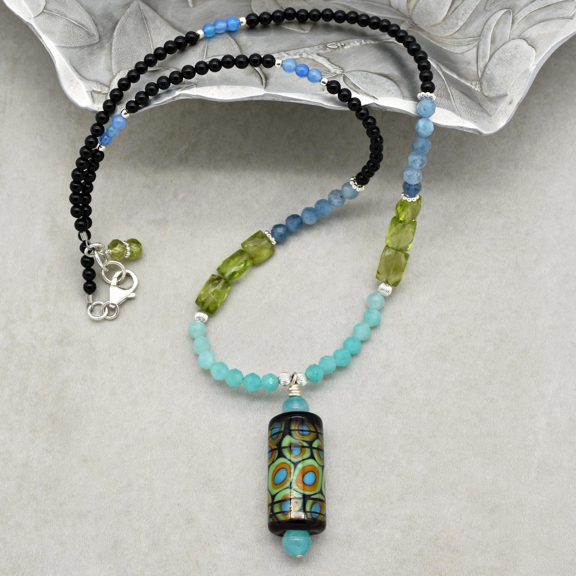 This image shows a necklace. The jewelry piece consists of various colored beads, including black, blue, green, and turquoise, with a large multicolored central pendant hanging in the middle. It also has a clasp, which is typical for necklaces.