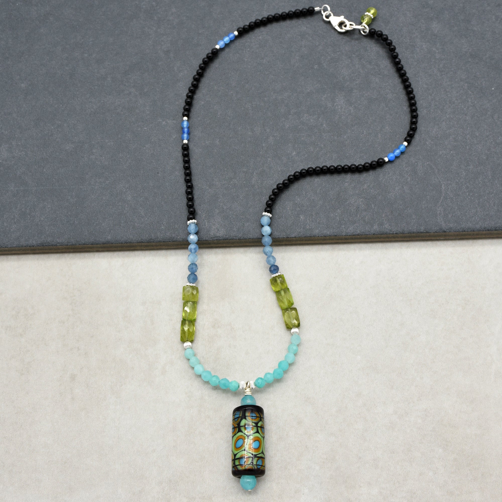 This image shows a necklace. The length and the clasp design, along with the pendant hanging at the center, are characteristic features of a necklace. It is made with a variety of beads in blue, green, and black tones.