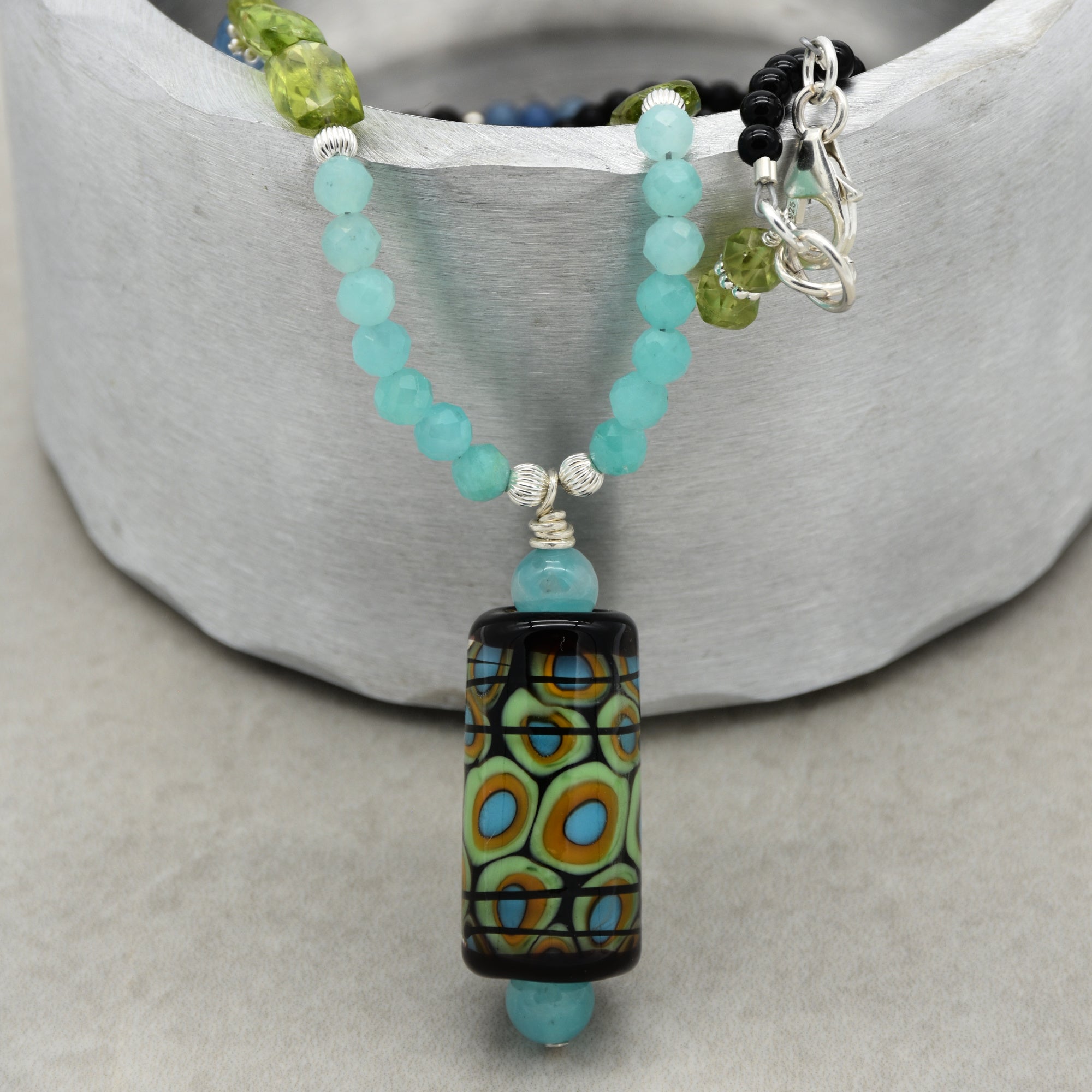 This image shows a necklace. The jewelry piece has turquoise beads, green beads, and a large decorative pendant with colorful patterns hanging in the center. The clasp and chain are also visible on the right side, typical of a necklace.