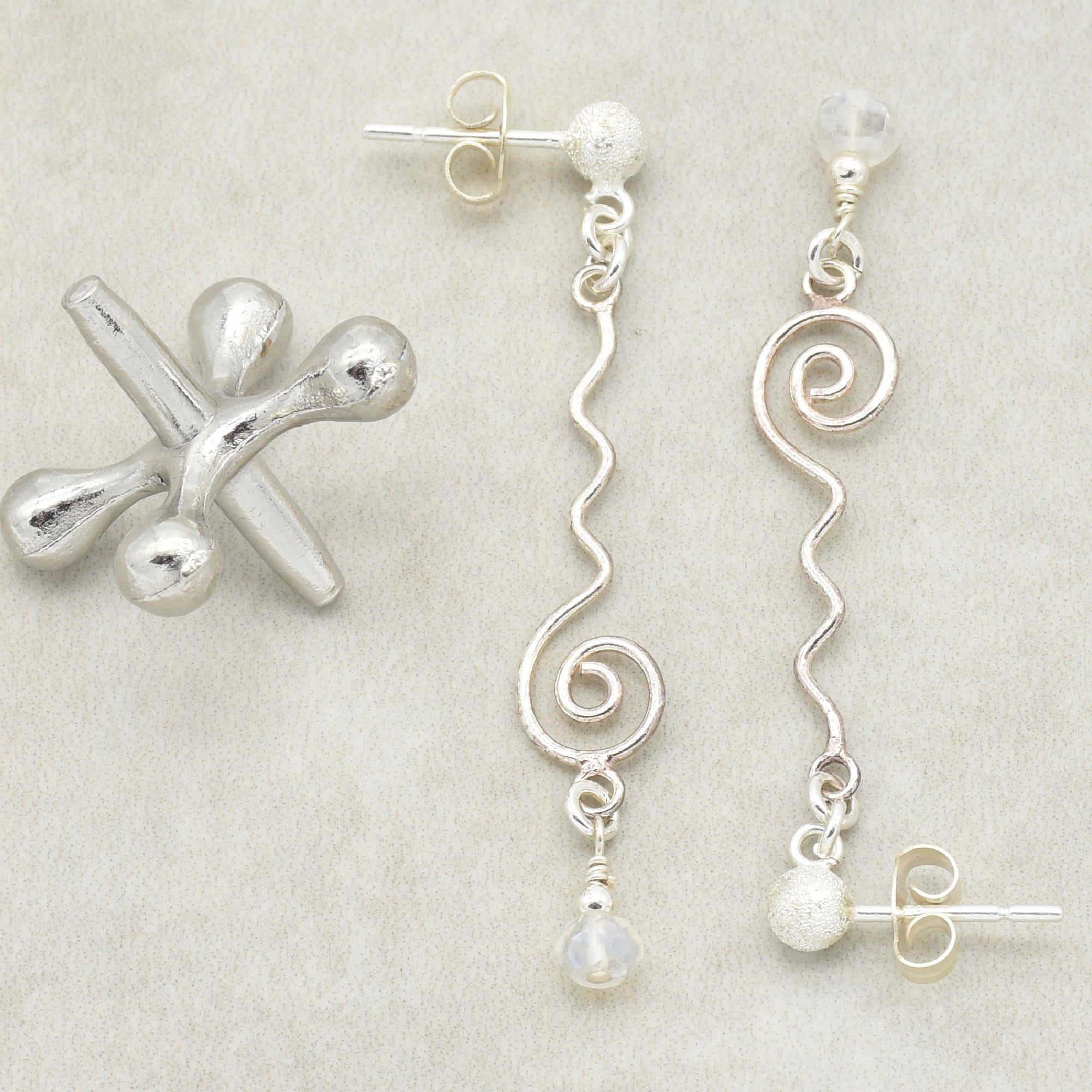 The image shows a pair of earrings. The earrings have a wavy, spiral silver design with a small bead hanging at the bottom and a stud backing for pierced ears.