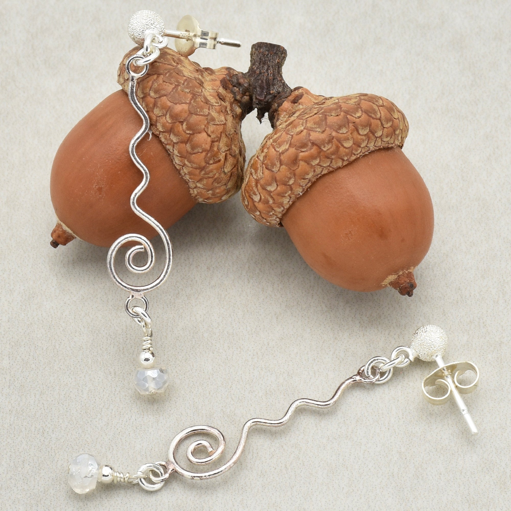 This image shows a pair of earrings. They have a silver, wavy, and spiral design with a small bead hanging at the bottom. The earrings are displayed with two acorns.