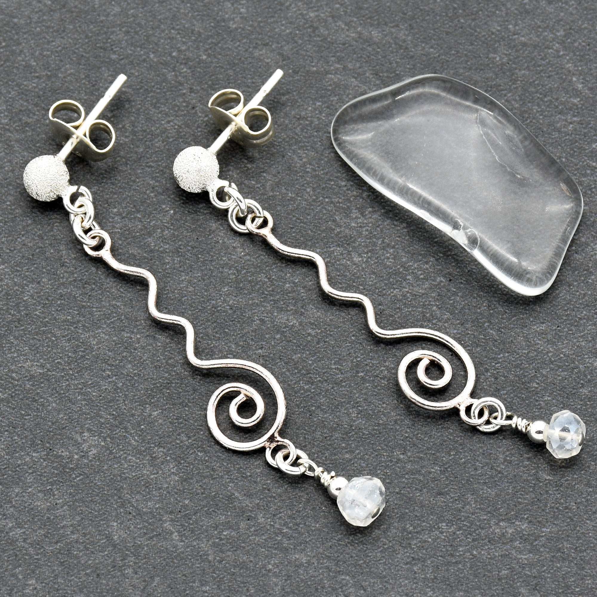 This image shows a pair of earrings. They have a post back design with a wavy silver wire and a small clear bead at the end.