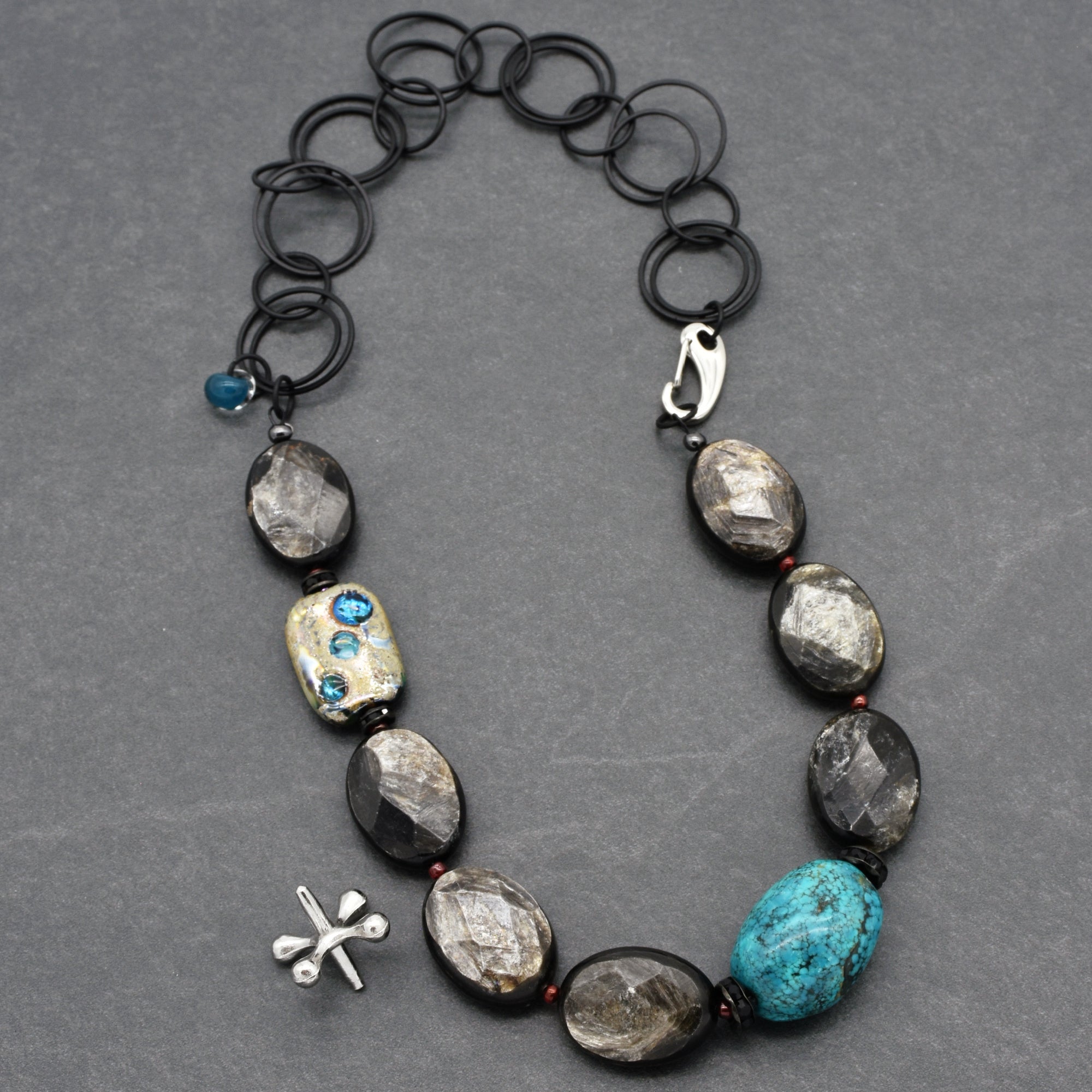 This image shows a necklace composed of alternating larger oval stones, mostly in a dark grayish color with one prominent turquoise stone, connected by a series of linked circular black rings near the clasp. There is also a silver dragonfly-shaped charm attached near the stones.