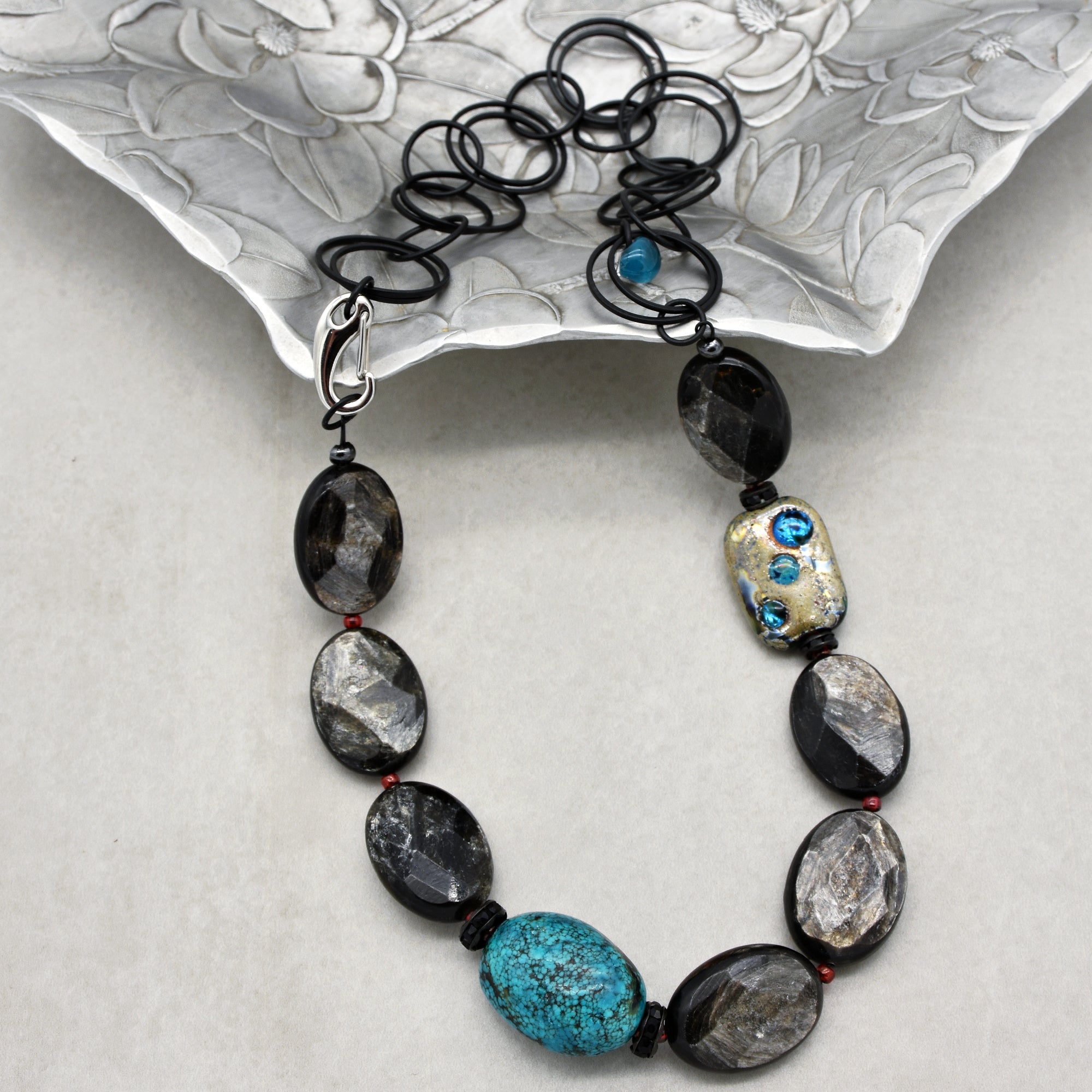 This image shows a necklace. It features a chain of large, oval-shaped black and silver-toned beads alternating with a prominent turquoise bead. The necklace also has a distinctive black looped chain section with a silver clasp for fastening. The combination of materials and colors gives it an eye-catching and elegant look.
