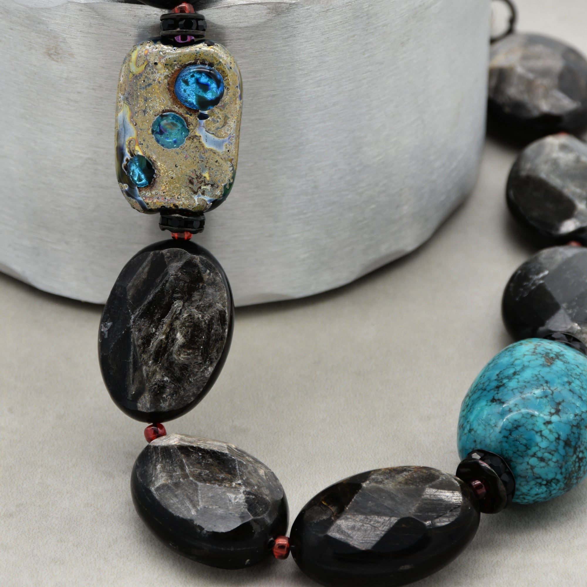 The image shows a close-up of a necklace. This can be identified by the arrangement and size of the beads, which typically are larger and strung together in a manner consistent with necklaces. The beads include a distinctive rectangular bead with blue and gold colors, as well as various textured oval beads in shades of black and turquoise.