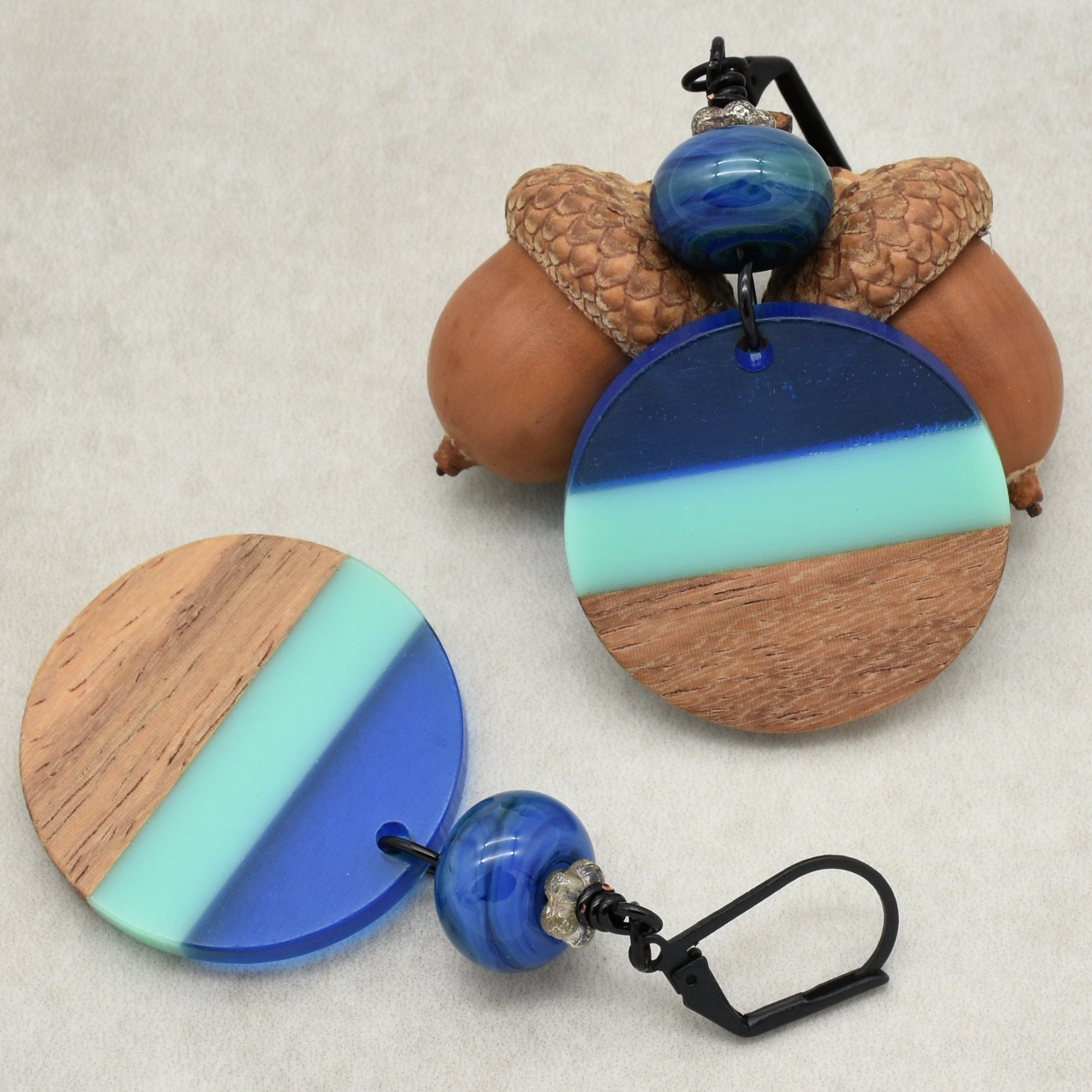 The image shows a pair of earrings. They have circular pendants with a design featuring wood and colored sections in blue and mint green. The earrings also have blue beads above the circular pendants and black metal hooks for wearing.