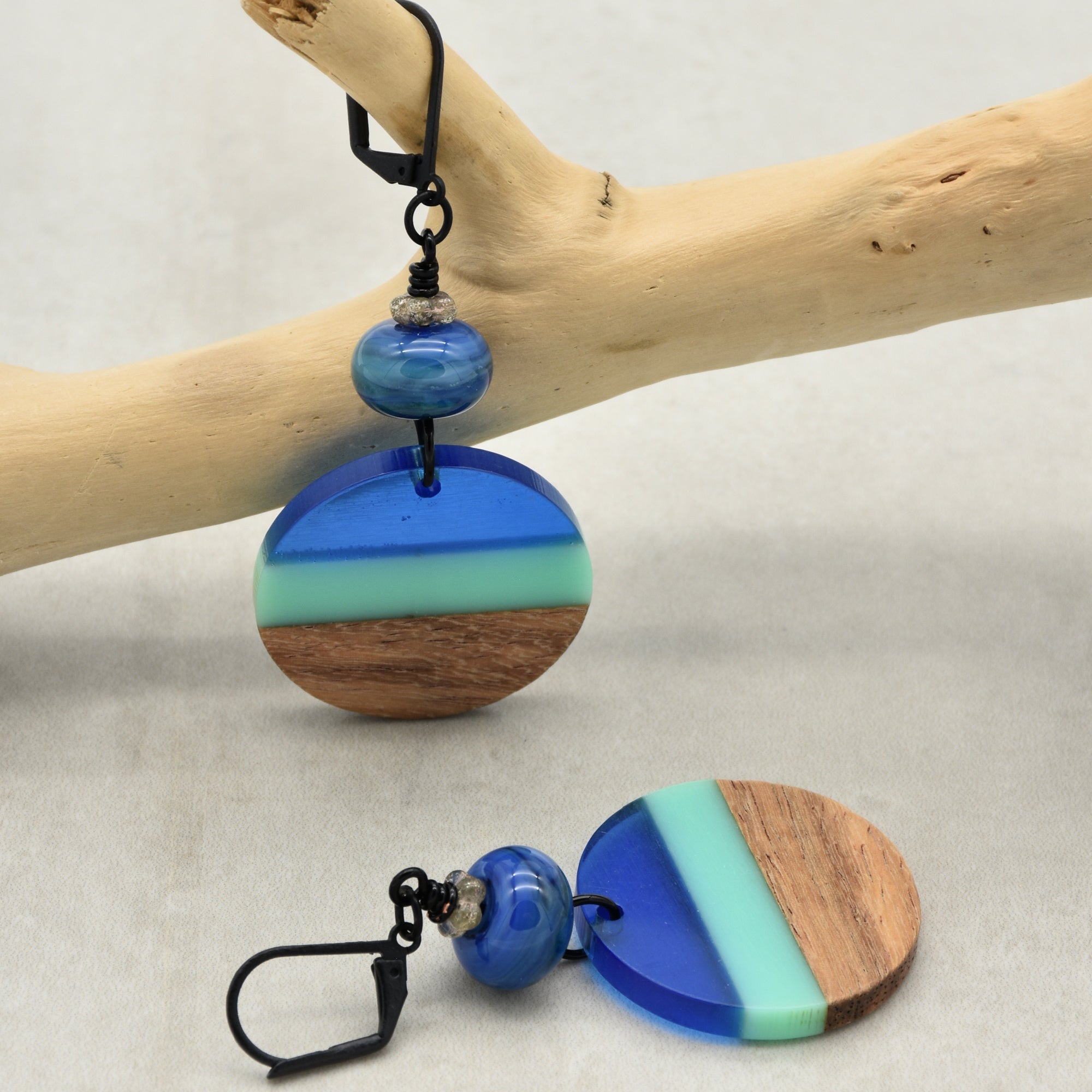 The image shows a pair of earrings. The earrings have a round, segmented design featuring blue, light green, and wood-toned colors, along with a small blue bead above the round piece. They are hanging from black hooks.