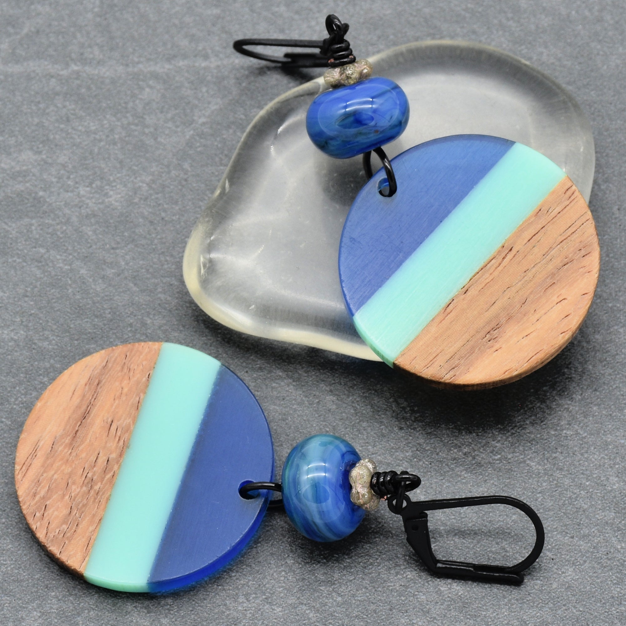 This image shows a pair of earrings. The jewelry pieces have black ear hooks and feature circular dangle elements with a tri-color design in blue, light green, and wood tones.