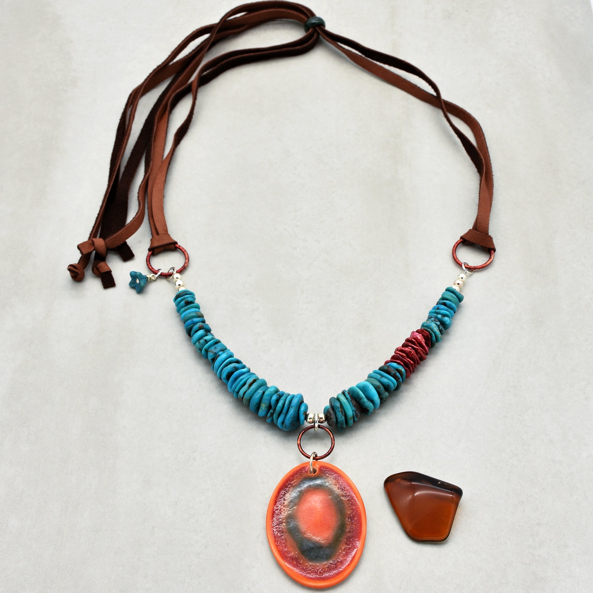 This image shows a necklace. It has a string of blue and red beads and a large oval orange pendant hanging in the center. The necklace is tied with brown leather straps.