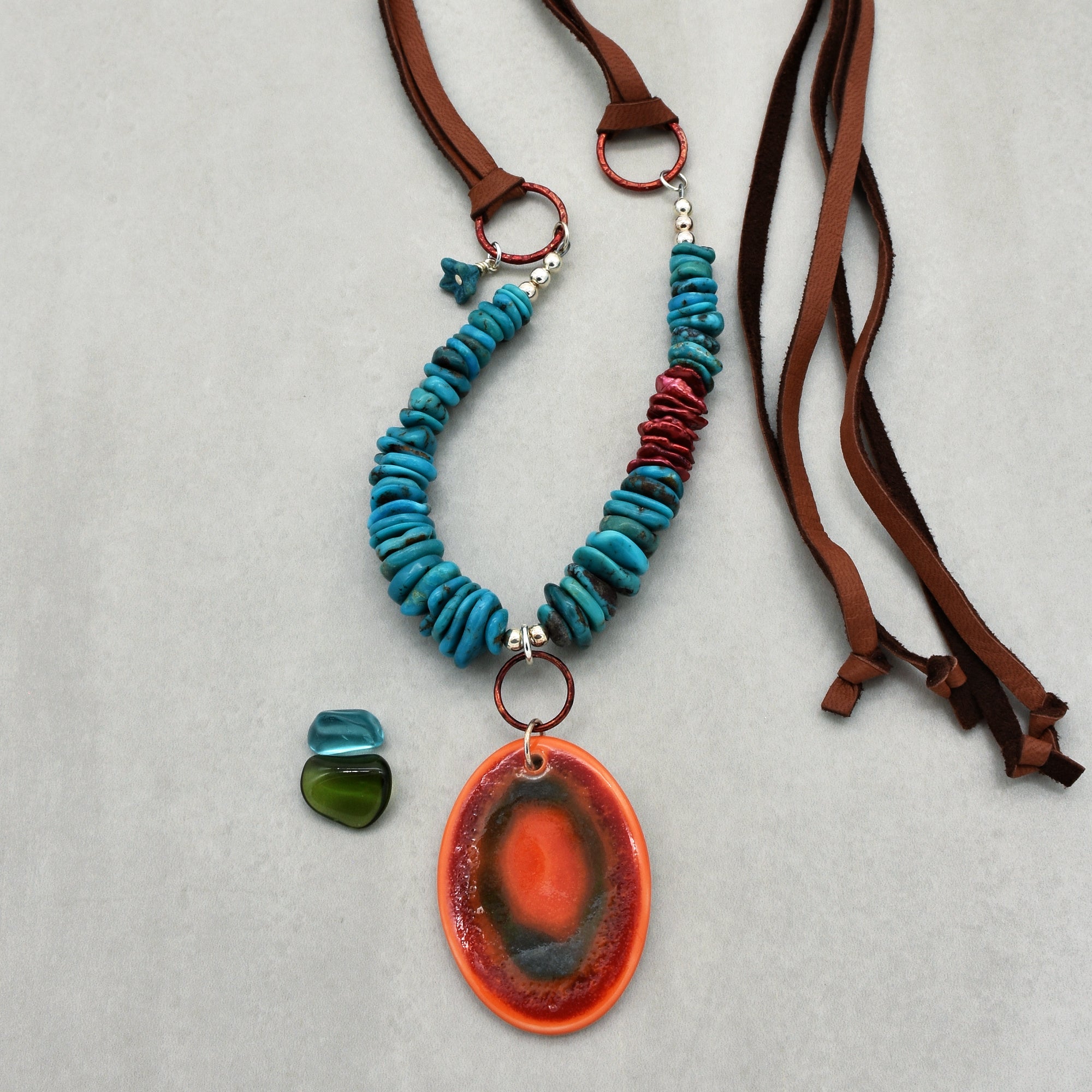 This image shows a necklace. It features blue beads and a large orange and black oval pendant, strung on a brown leather cord.