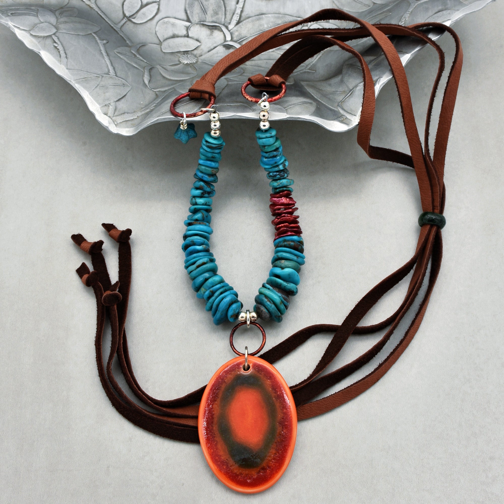 This image shows a necklace. It features turquoise stone beads and a large oval pendant with a reddish-orange and dark center, strung on brown leather or suede cords.