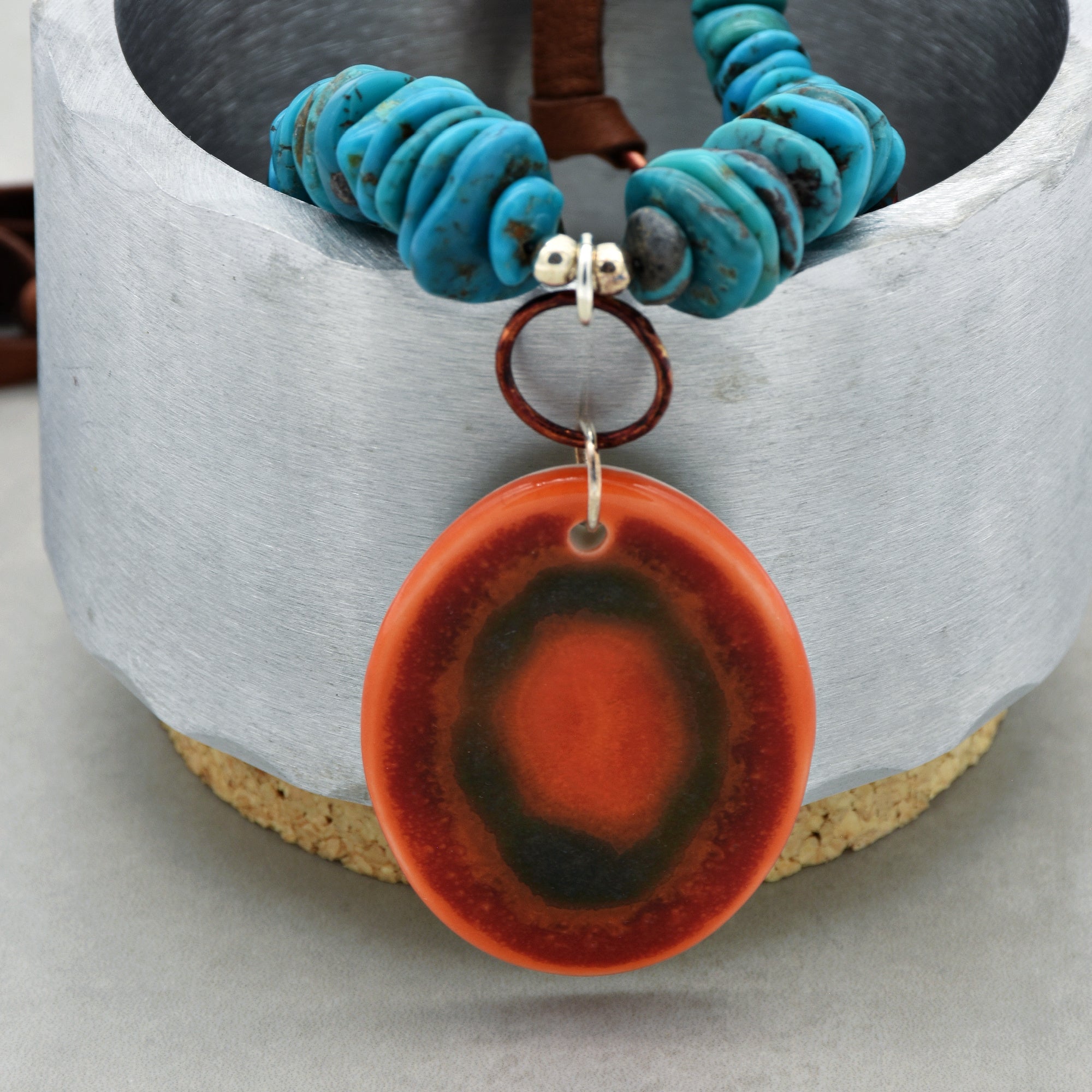 This image shows a necklace. The jewelry consists of blue turquoise beads and a large, round orange and dark brown pendant hanging from the center.