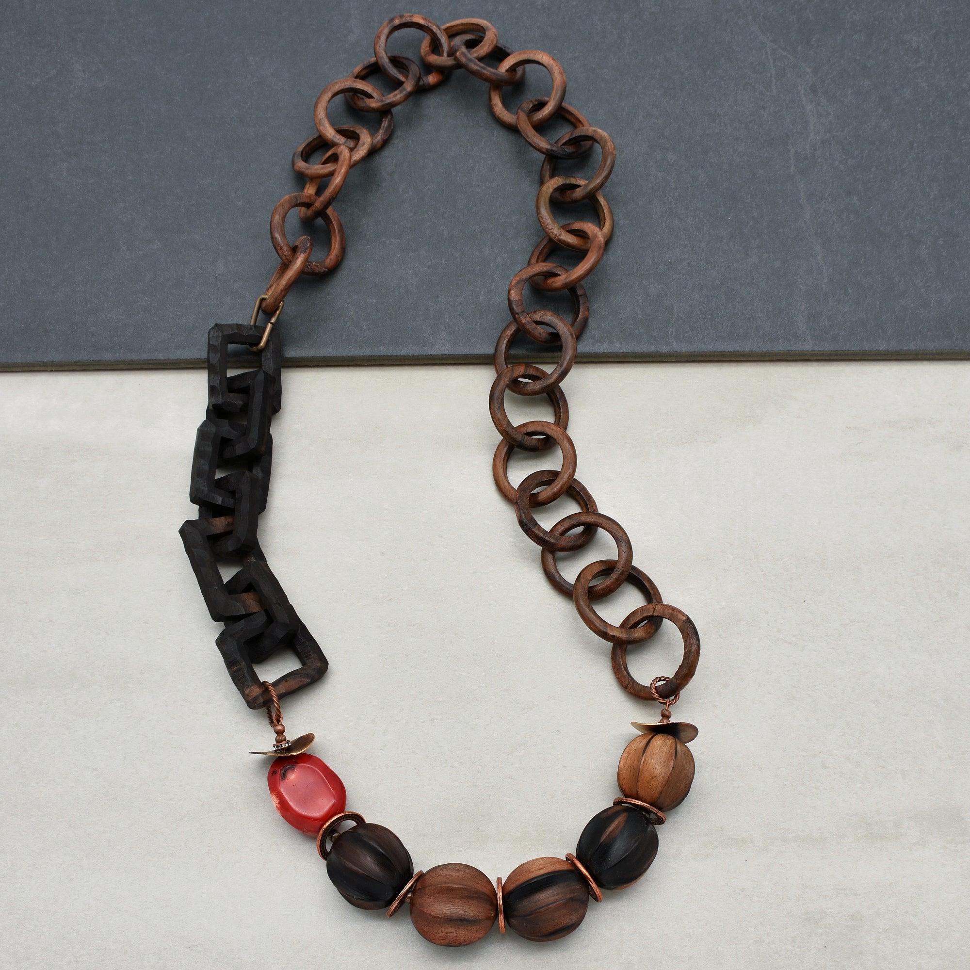 This image shows a necklace. The design includes large wooden chain links and several round beads, with one red bead among the wooden ones, arranged to form the piece of jewelry.