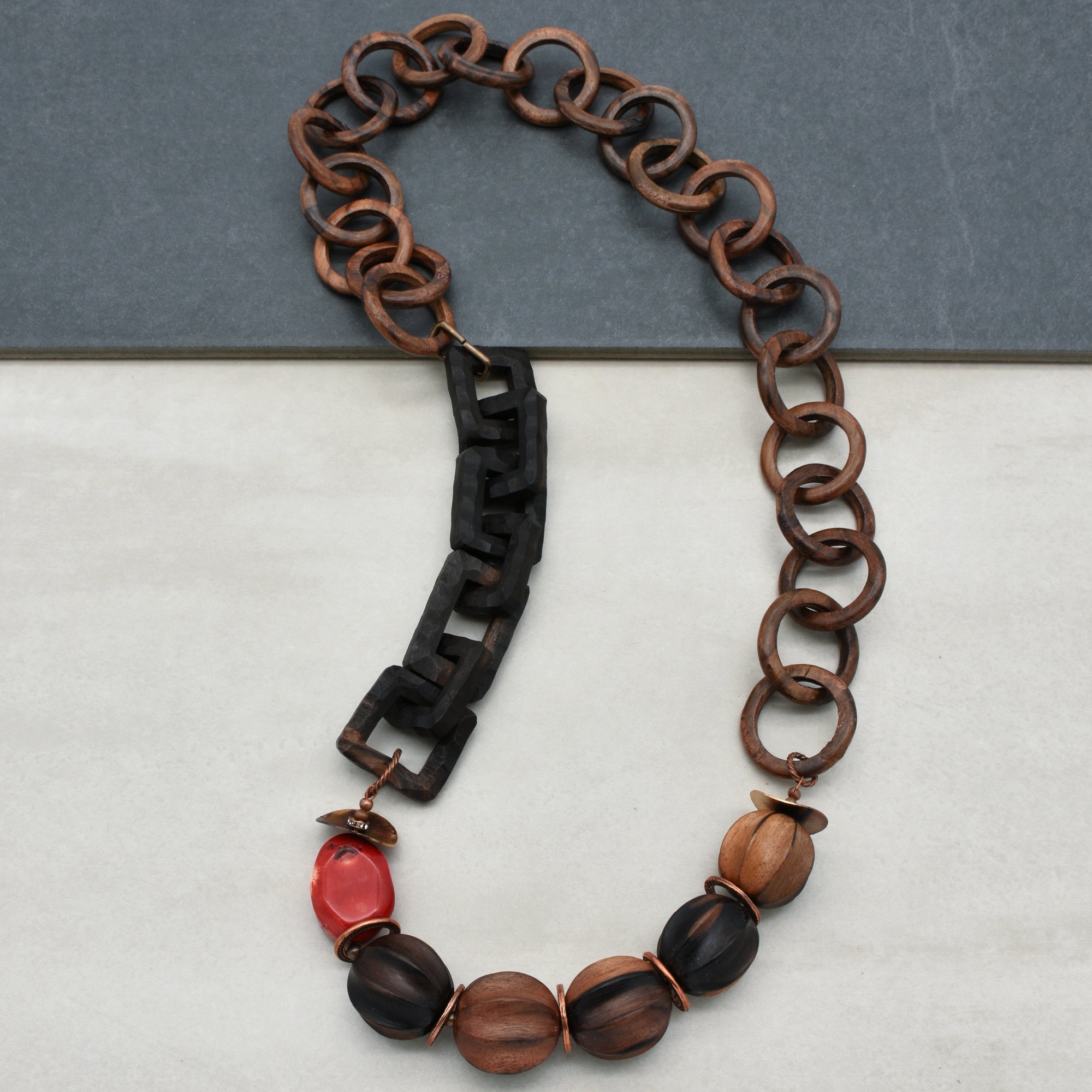 This image shows a necklace. It has large interlocking wooden links and a series of unique beads towards the bottom, including one that is red and several that appear to be carved wood. The style and length are consistent with a necklace rather than an earring or bracelet.