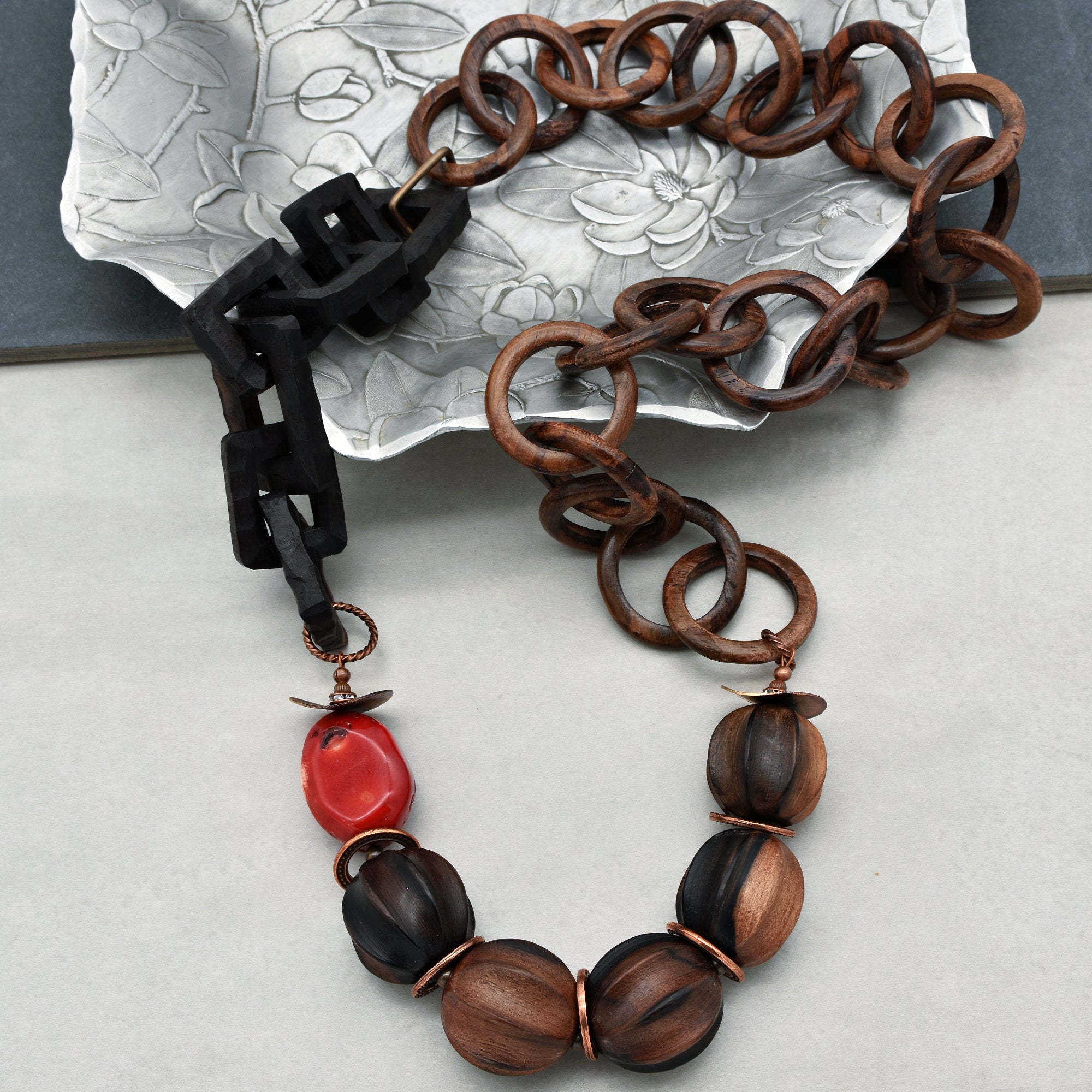 This image shows a necklace. The jewelry features a chain made of linked wooden rings and includes round wooden beads along with a single red bead as part of its design. The necklace has a mix of textures and colors, giving it an artistic and natural look.